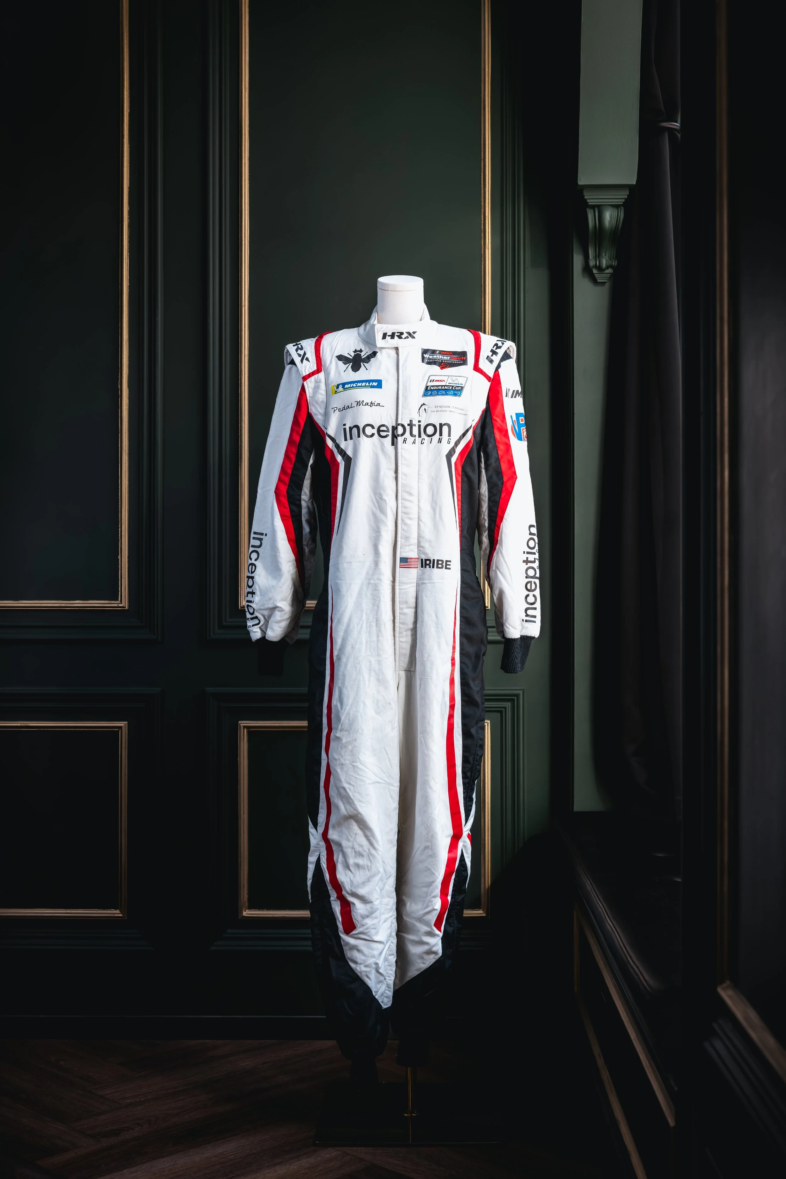 Racing suit on display, white with red and black accents, covered in sponsor logos, on a mannequin against a dark green paneled wall.