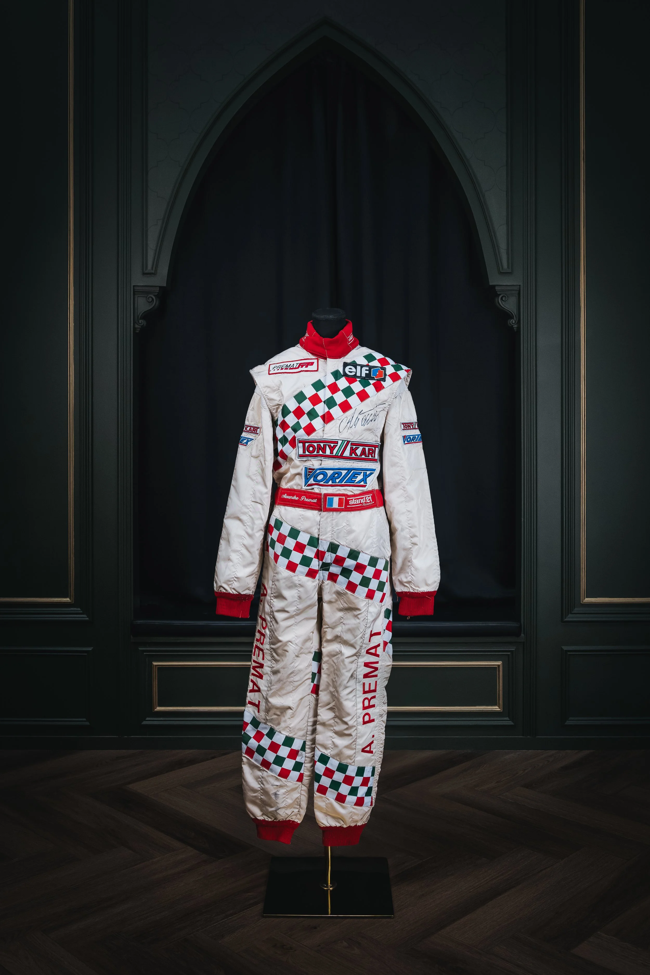 Racing suit with sponsor patches and checkered pattern, displayed on a mannequin against a dark background.
