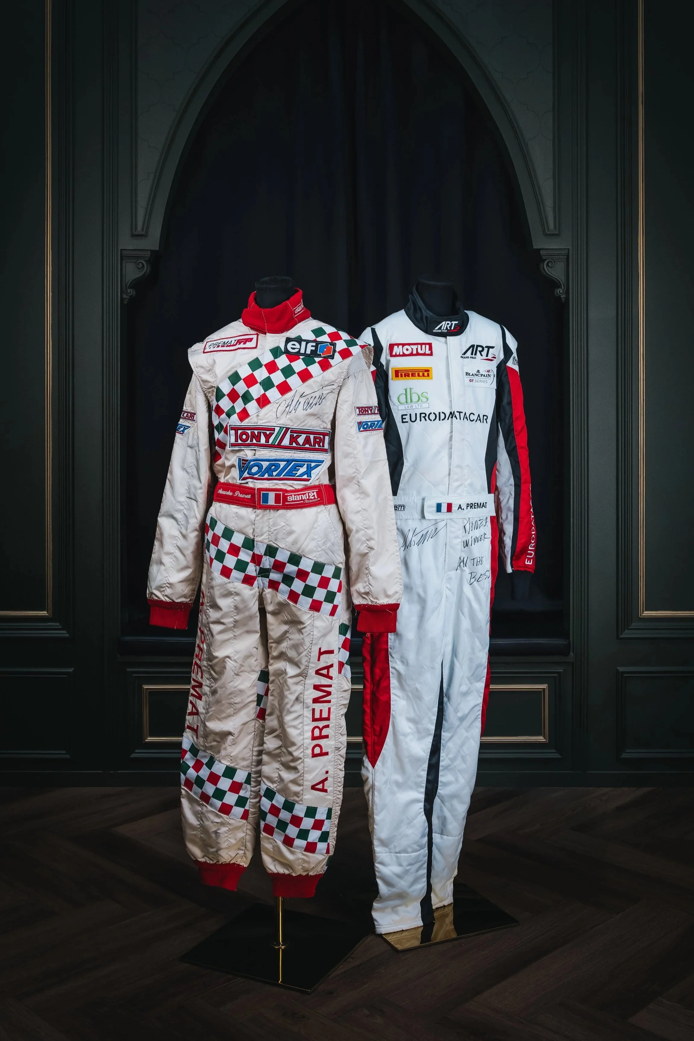 Two racing suits on mannequins inside a display case. The left suit is mainly white with red, green, and black checkered patterns, and the right suit is mainly white with red and black accents. Both suits have various sponsor logos and signatures.