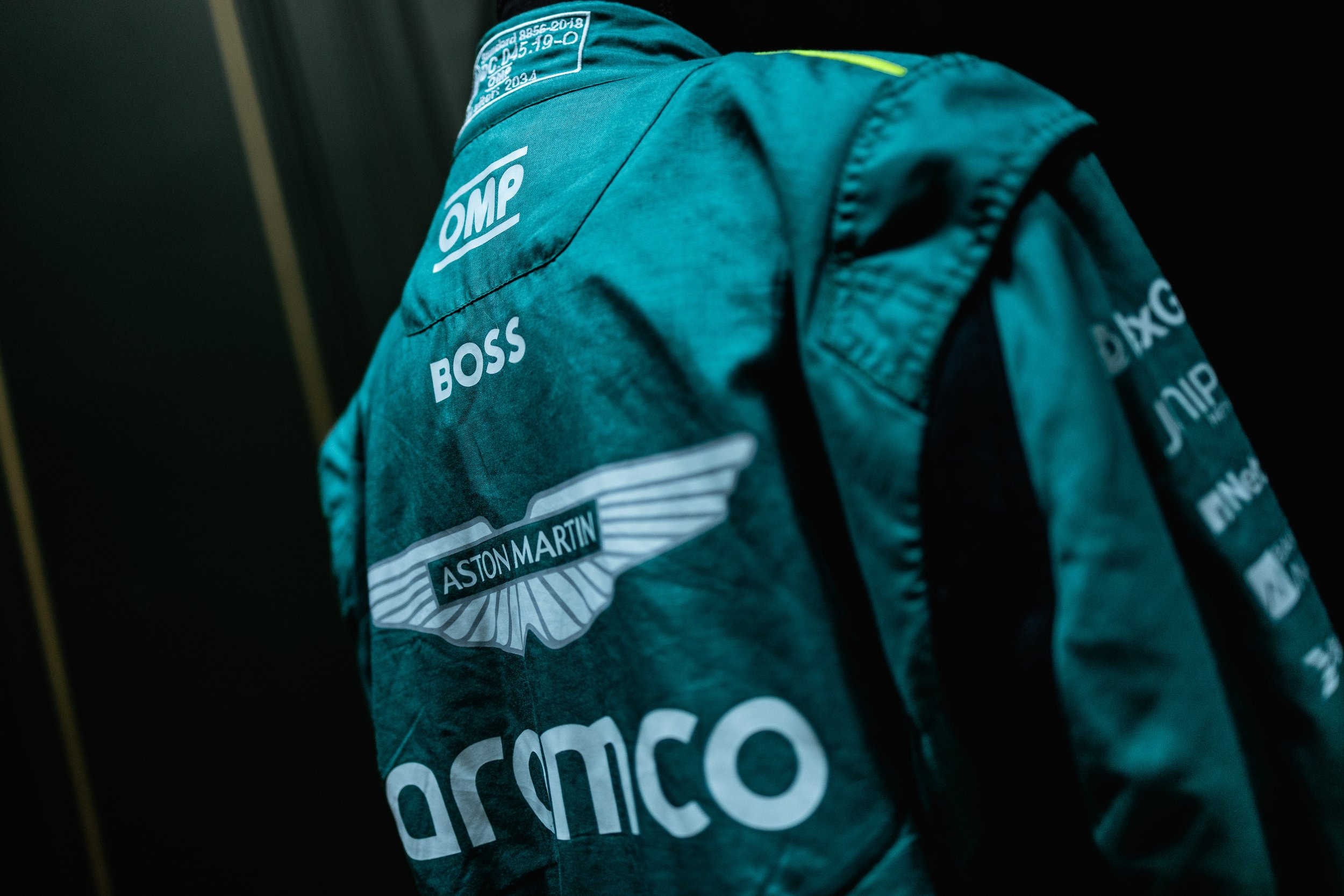 Close-up of a teal racing suit with various sponsor logos, including Aston Martin, OM, and Aramco, and the word 'BOSS' on the back.