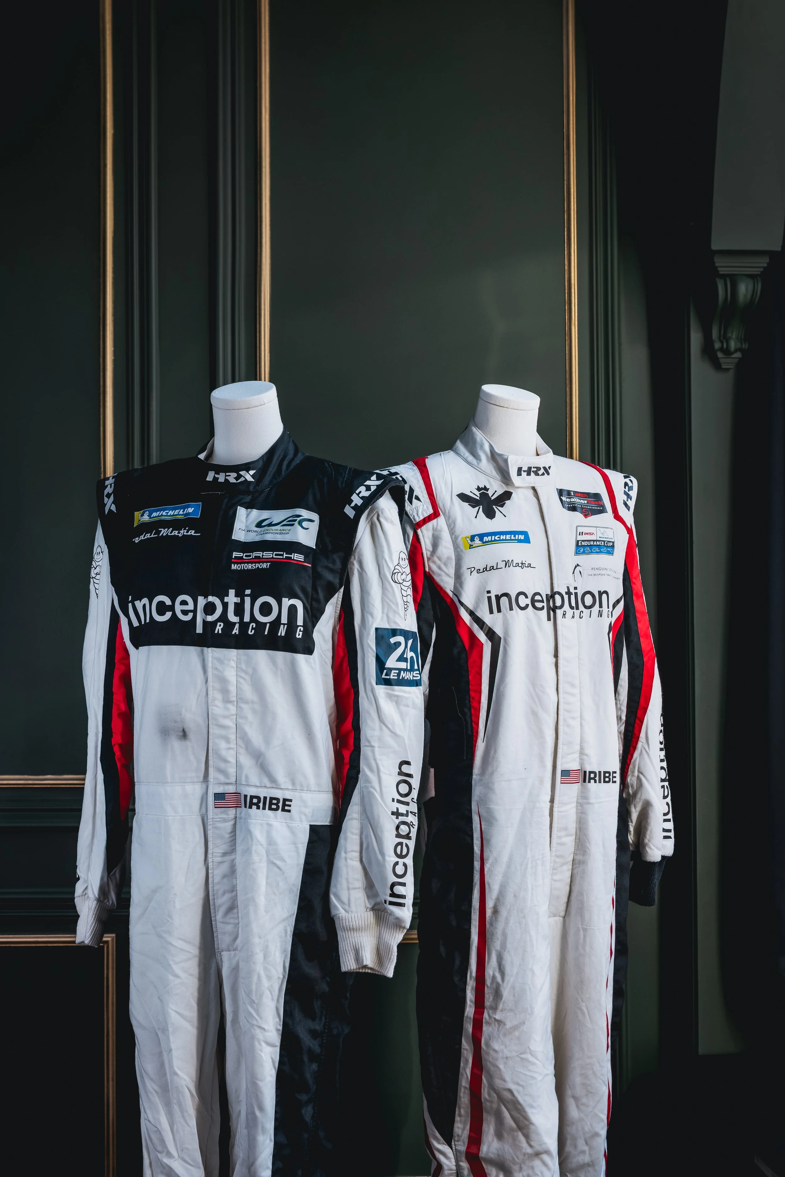 Two racing suits on mannequins, one black and white with logos, the other white with red and black accents, displayed against a dark green wall.