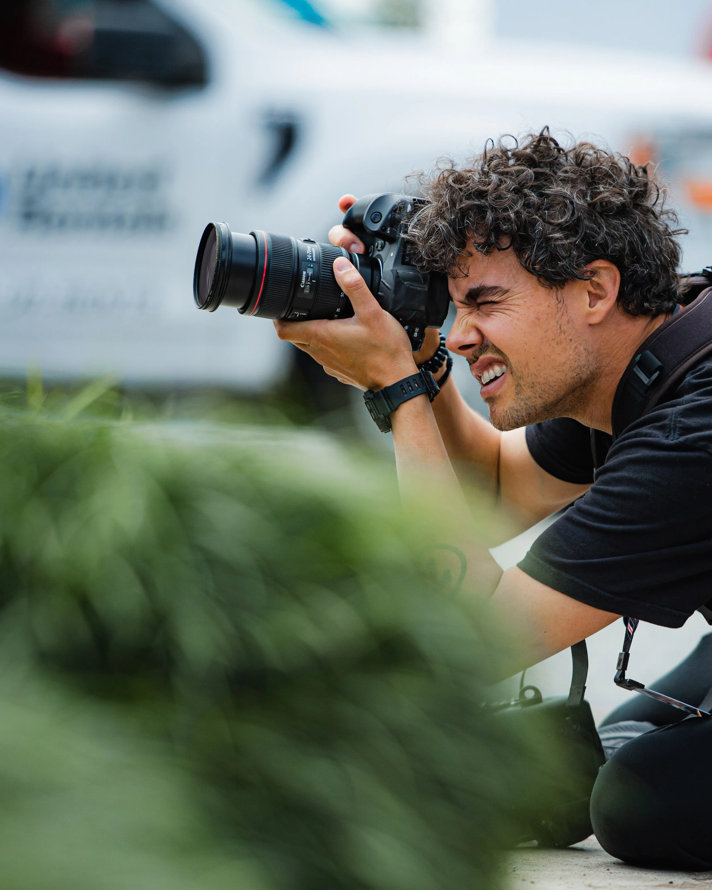 A man with curly hair taking a photo with a professional camera, crouched down on the ground.