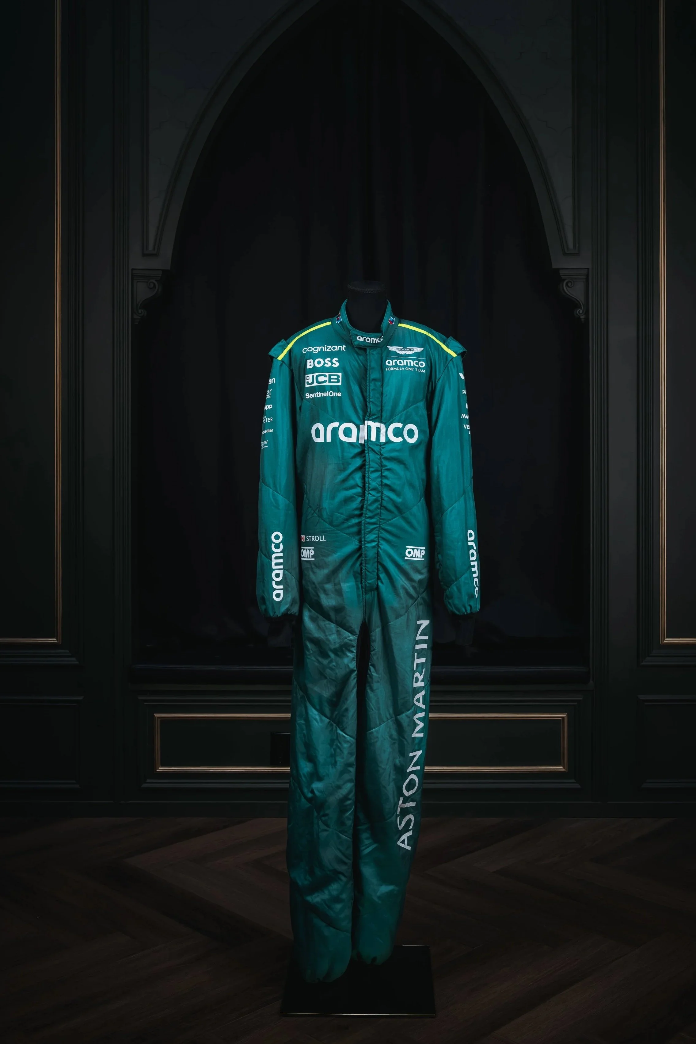 A racing suit displayed on a black mannequin in front of a dark, ornate wooden background. The suit is teal with various sponsor logos, including "Aramco" prominently on the chest and "Aston Martin" on the leg.