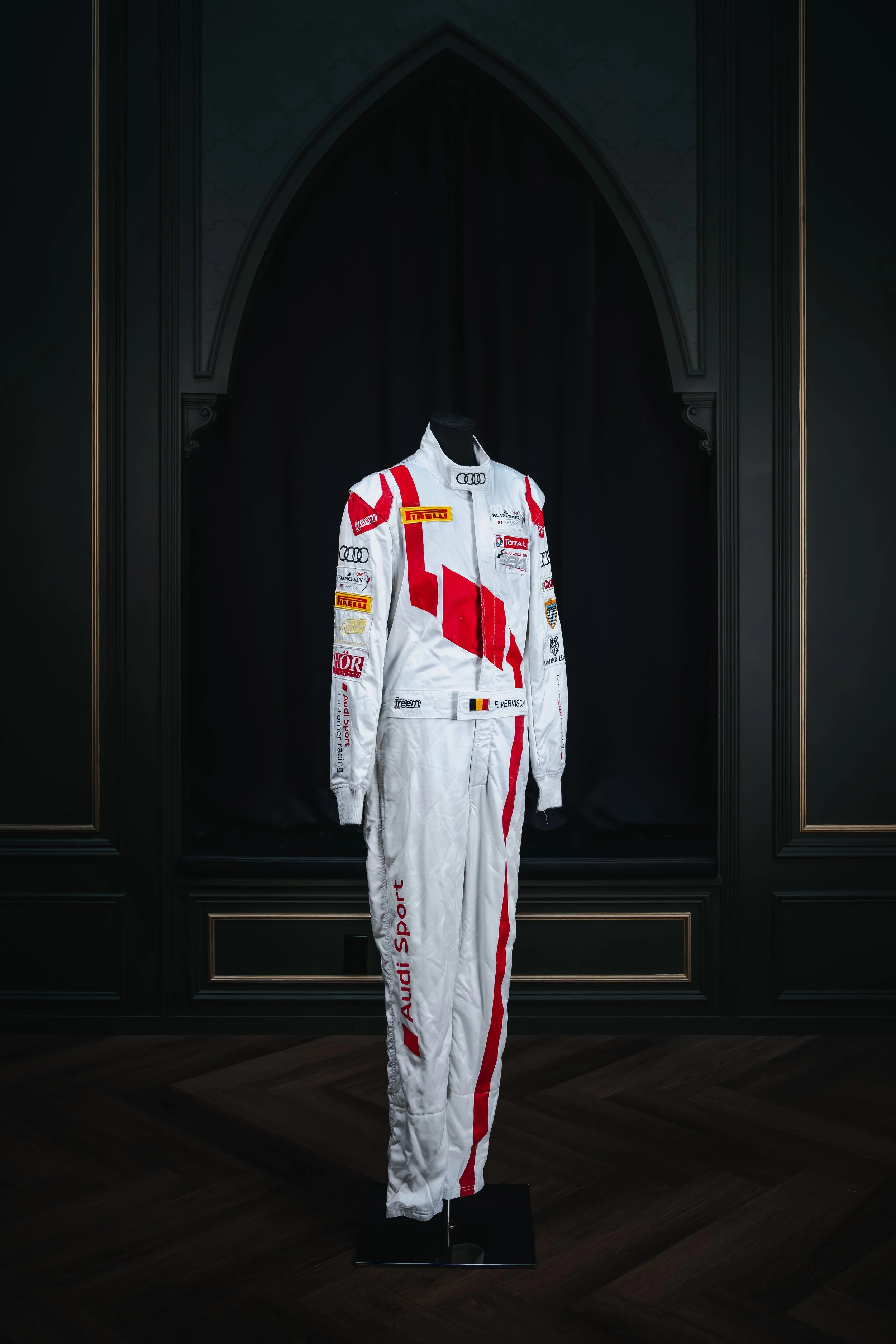 A race car driver’s white racing suit with red and black accents, displayed on a black mannequin against a dark background with a black curtain.