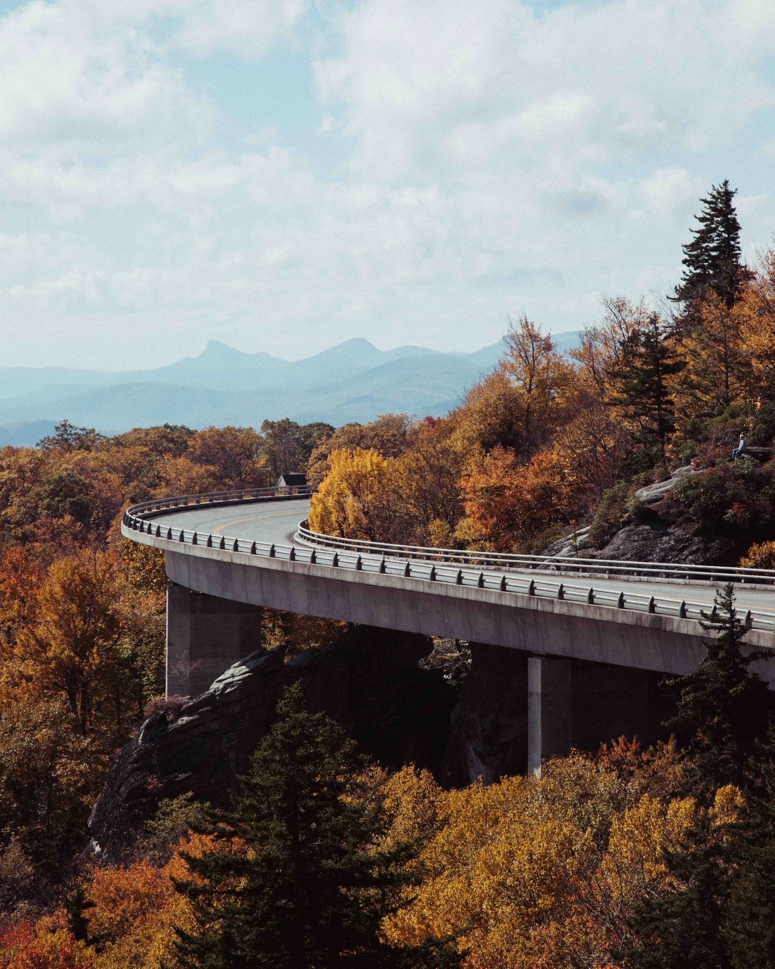 A scenic view of a curved highway bridge in a forested area with autumn-colored trees, mountains in the background, and a partly cloudy sky.
