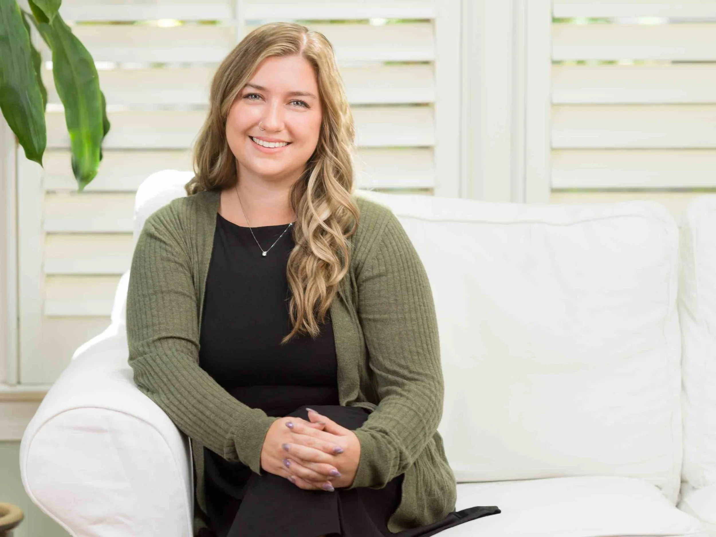 A woman with long, wavy blonde hair sitting on a white couch, smiling, in a bright room with white shutters and a plant in the background.
