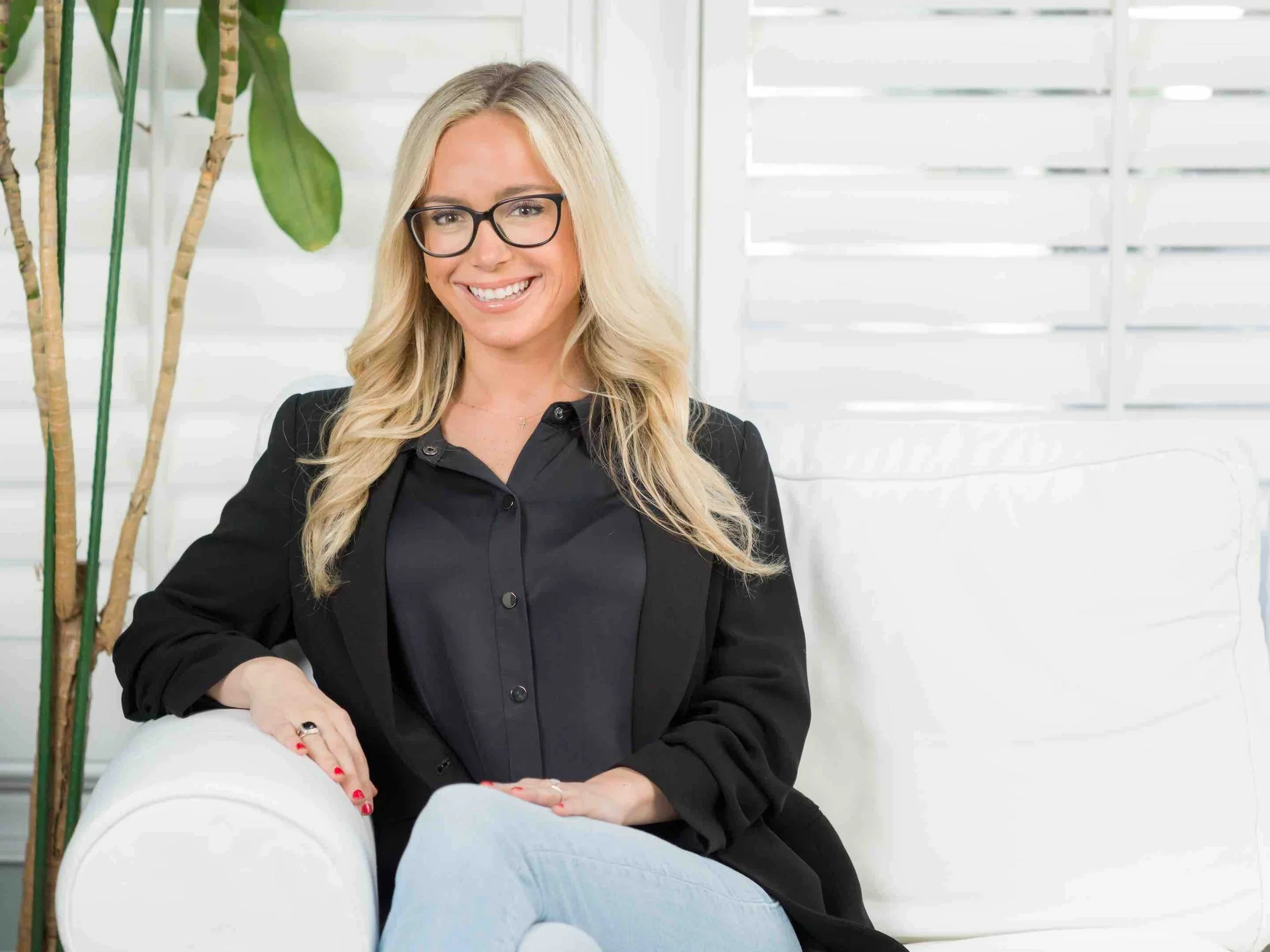 A woman with blonde hair, glasses, and a black blazer sitting on a white sofa, smiling in a bright room with a plant and white blinds in the background.