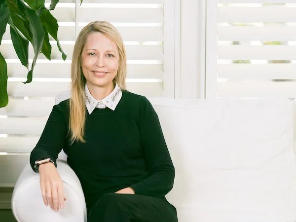 A woman with blonde hair sitting on a white couch, wearing a black dress and white collared shirt, in front of white blinds and a green plant.