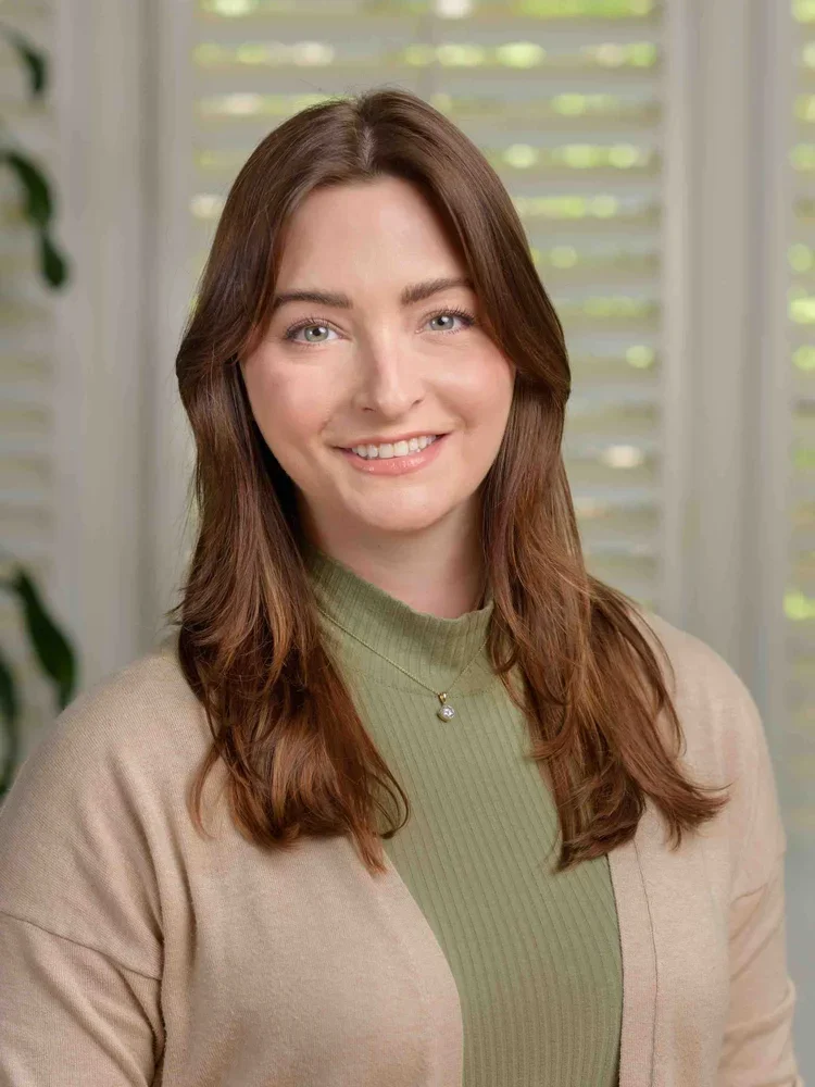 A woman with long brown hair and blue eyes, smiling, wearing a green ribbed turtleneck and beige cardigan, standing in front of a window with blinds.