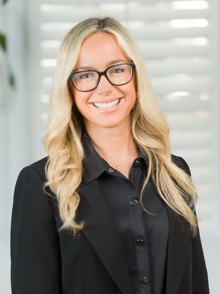 A smiling woman with blonde hair, wearing glasses and a black blazer, standing in front of a background with horizontal blinds.