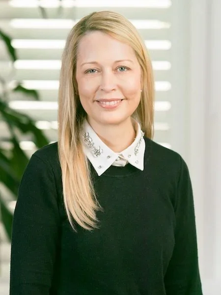 A woman with long blonde hair, wearing a black sweater and a white collared shirt with decorative details, smiling in front of a background with window blinds and green plants.