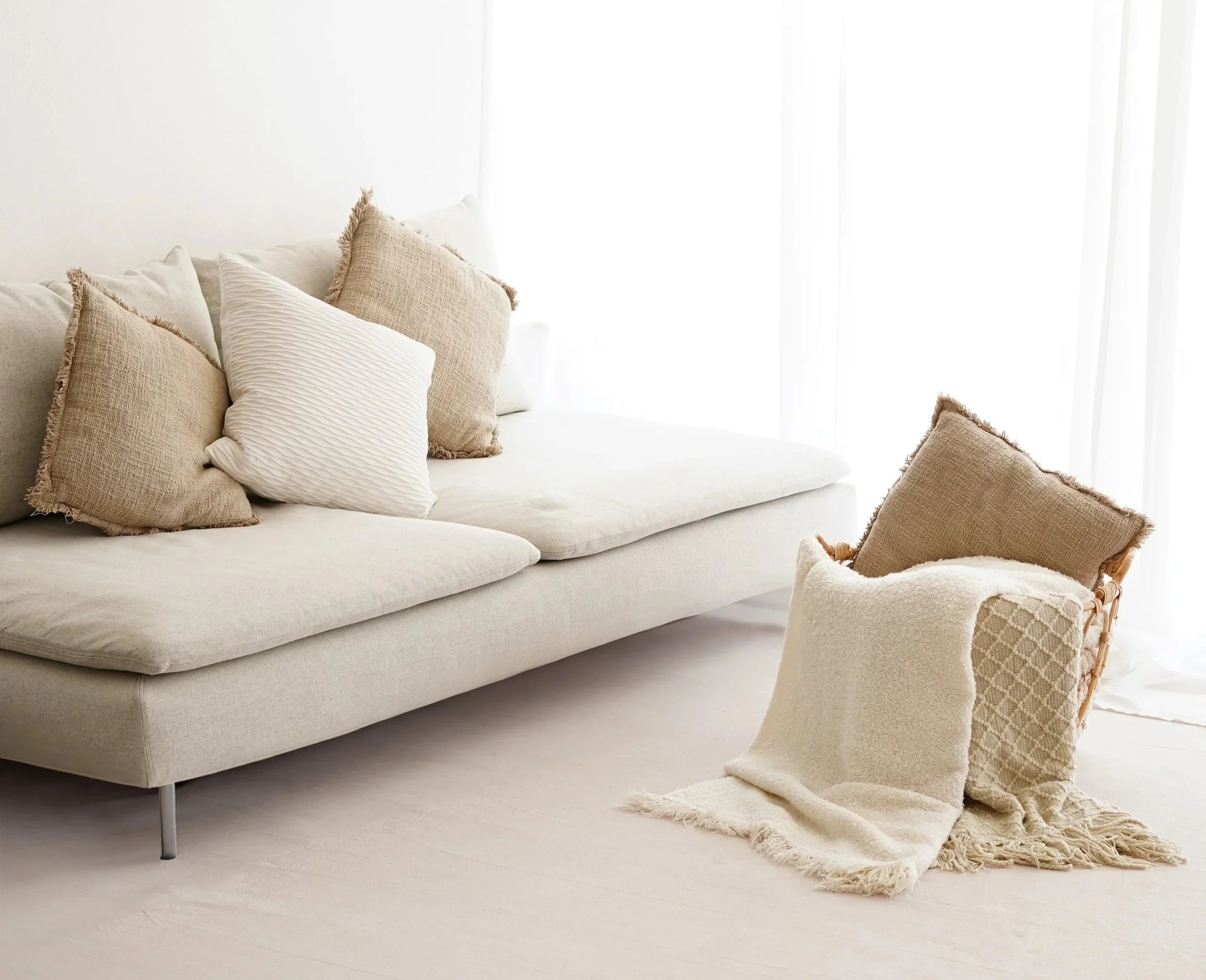 Ein helles Wohnzimmer mit einem cremefarbenen Sofa, mehreren Kissen in neutralen Tönen und einem Korbtuch mit einer Decke darauf, vor einem Fenster mit weißen Vorhängen.