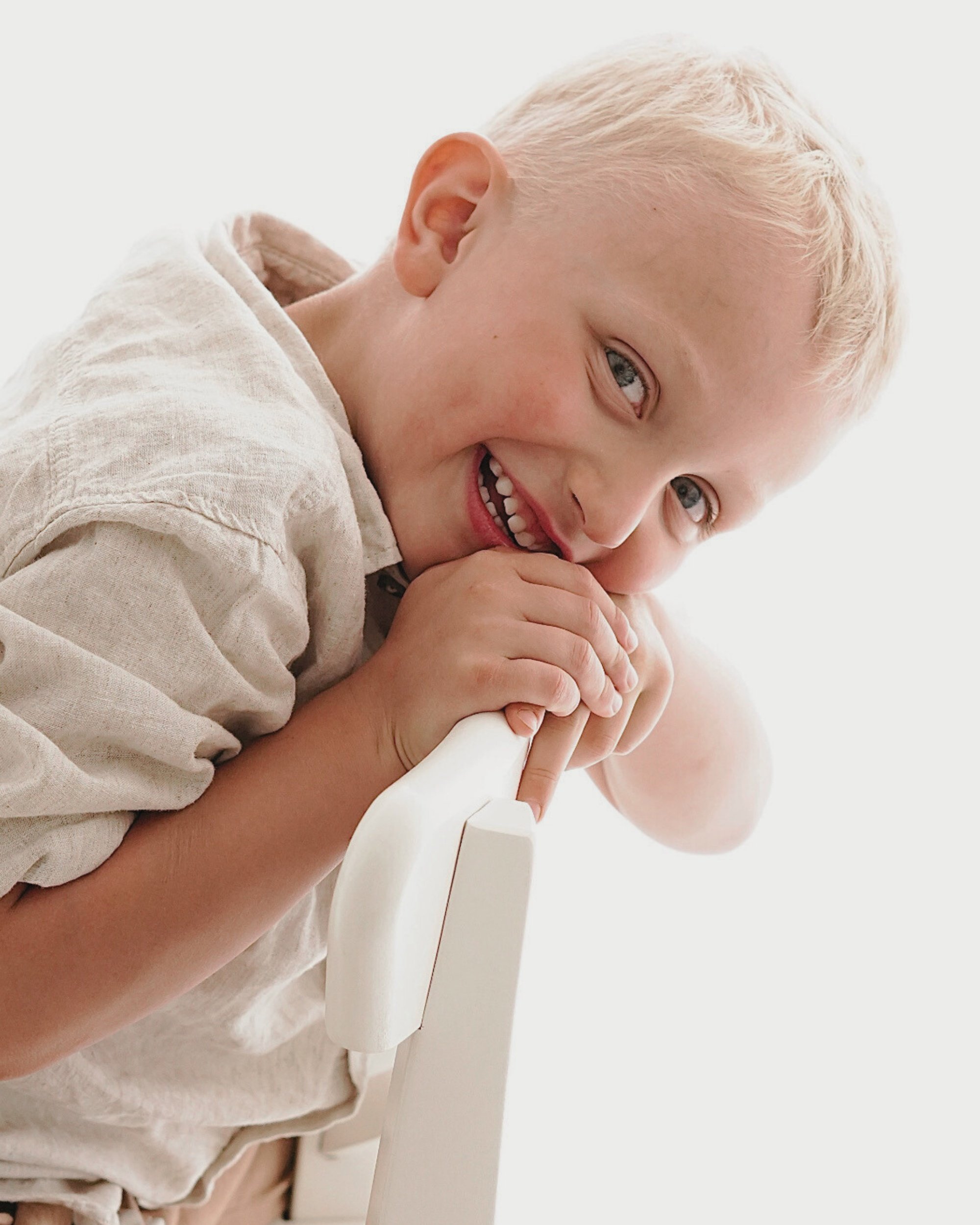 Ein lachender Junge mit blonden Haaren, der sich an einem weißen Geländer festhält.