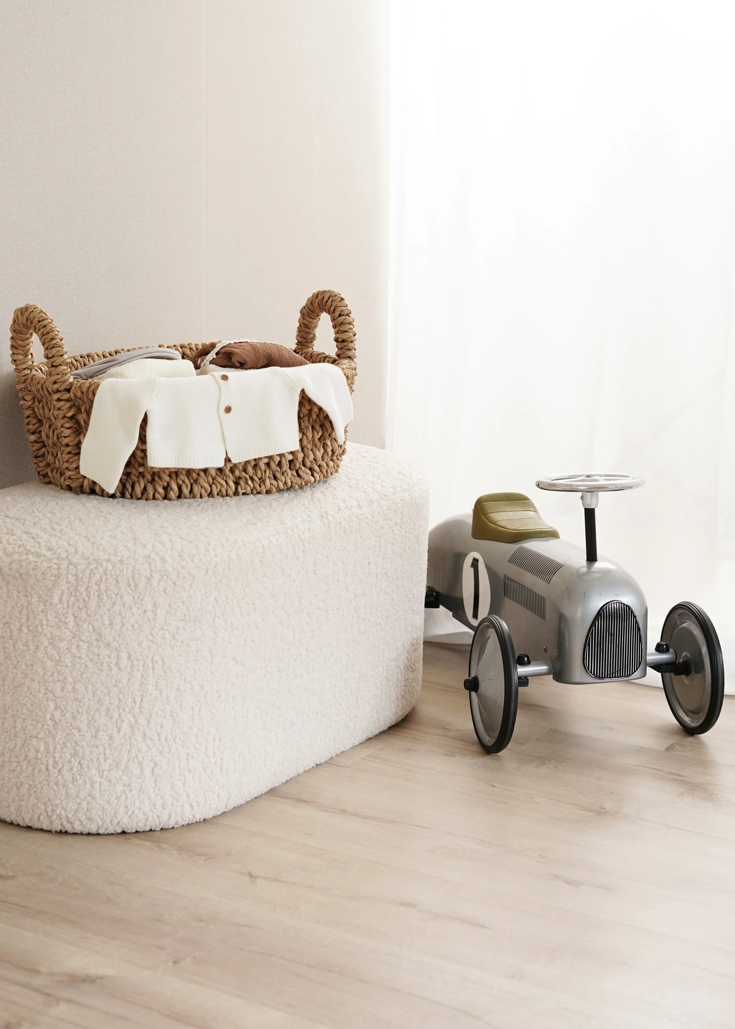 Ein Korb mit Babykleidung neben einem Kinderauto in einem Zimmer mit Laminatboden und weißen Vorhängen.