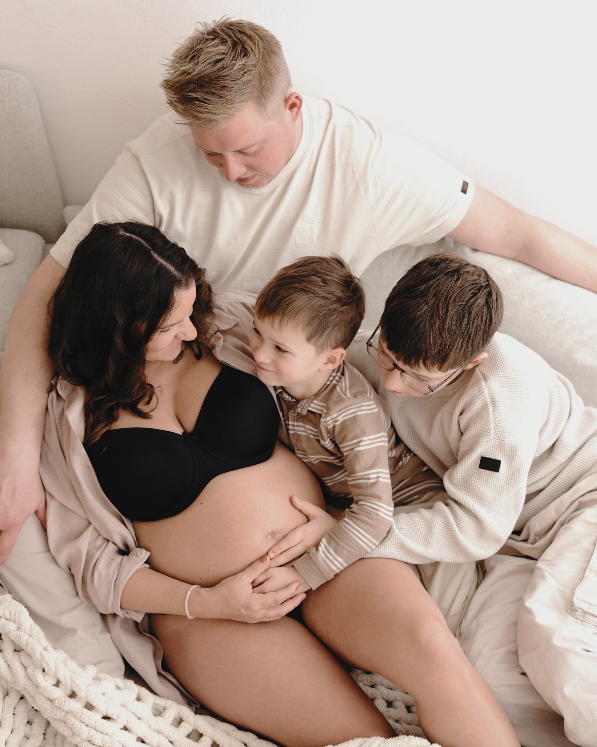 Eine schwangere Frau sitzt auf einem Bett, umgeben von einem Mann und zwei Kindern, die sie liebevoll berühren.