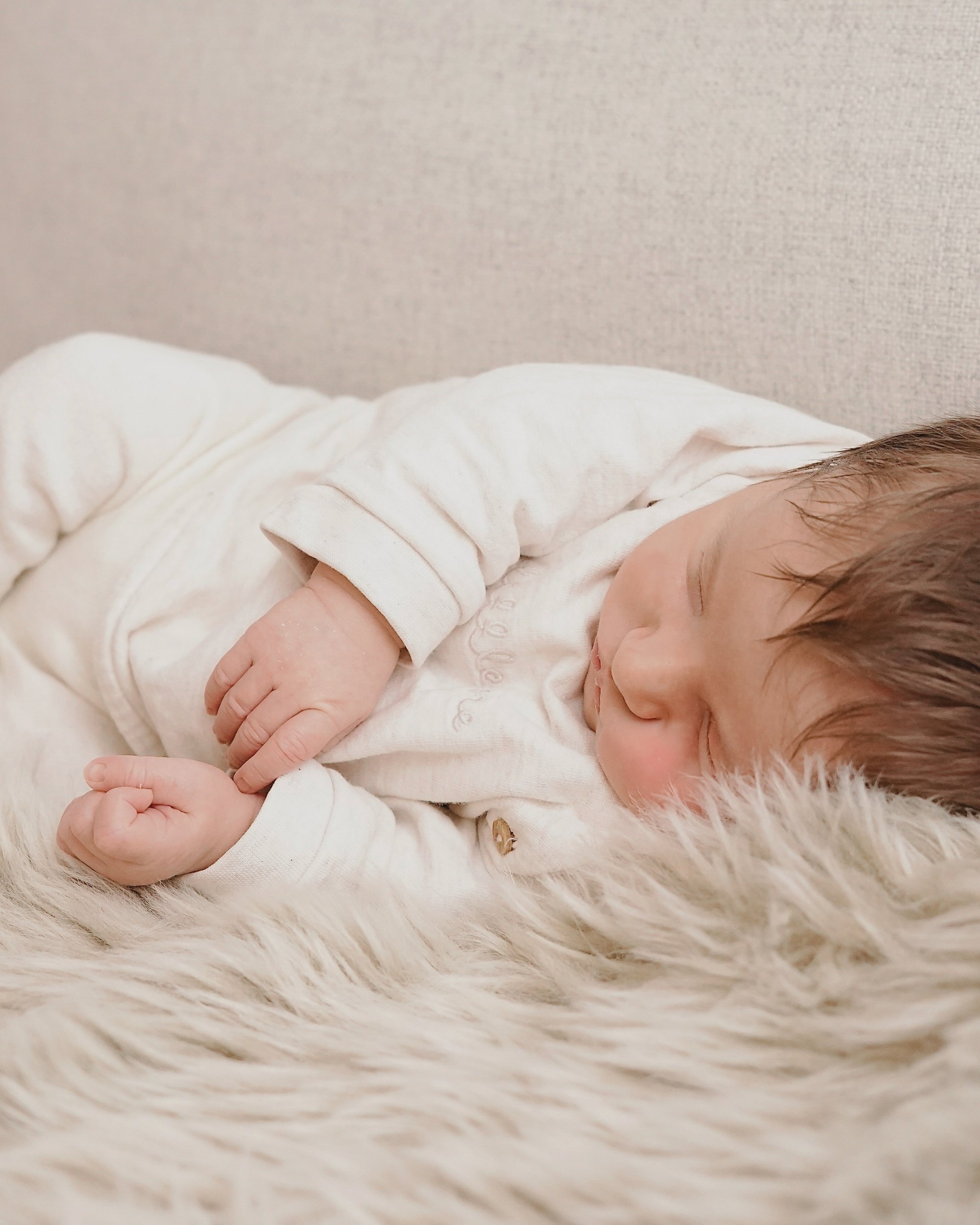 Ein schlafender Säugling liegt auf einer weichen, weißen Decke, eingekuschelt in einem weißen Outfit.