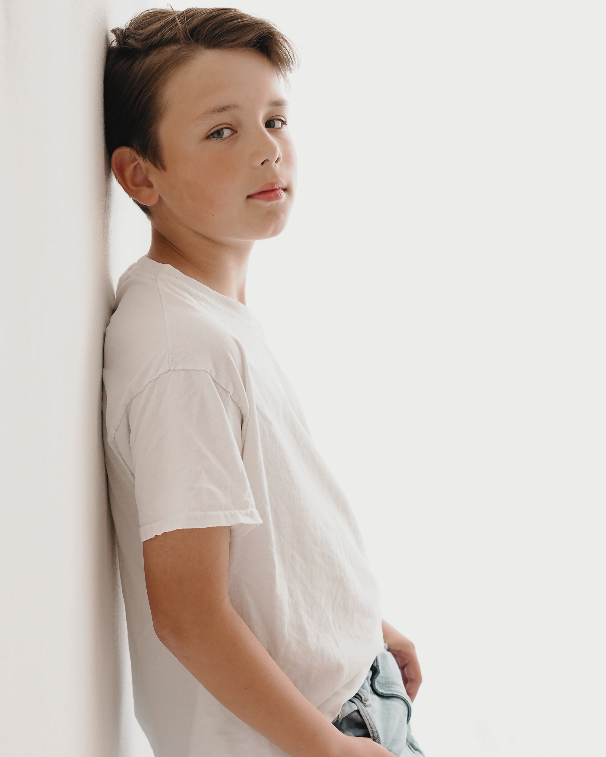 Junge steht an einer weißen Wand, trägt ein weißes T-Shirt, schaut in die Kamera, hat braune, kurze Haare.