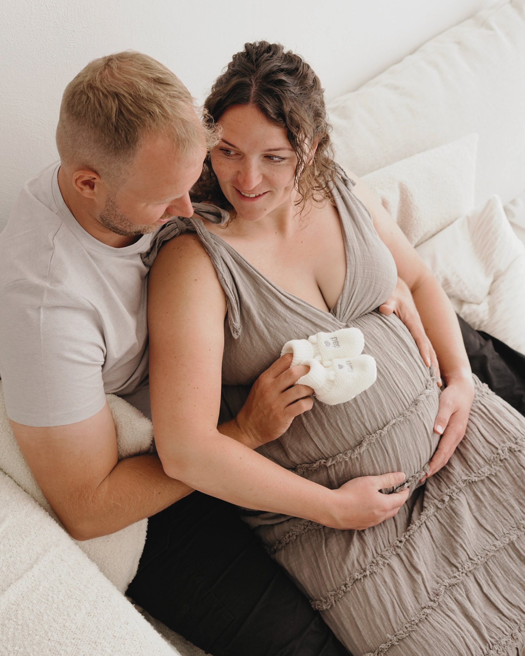 Ein werdendes Mutterpaar sitzt auf einer Couch, sie hält ihre schwangere Bauch und lädt kleine Babyschuhe auf ihren Bauch, während der Partner sie liebevoll umarmt.
