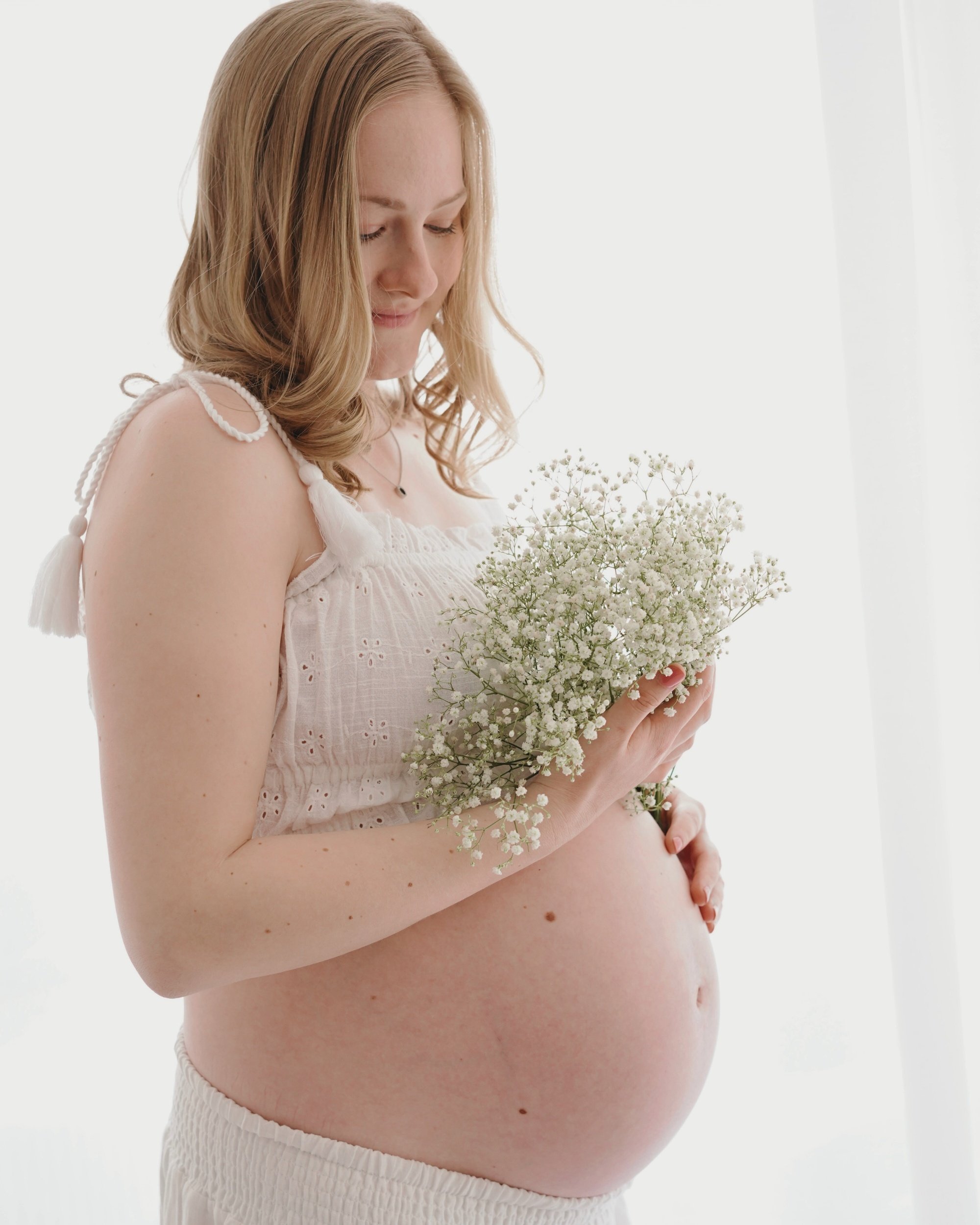 Eine schwangere Frau mit lockigem, hellbraunem Haar hält einen Blumenstrauß aus kleinen weißen Blumen vor ihrem Bauch, während sie nach unten blickt und lächelt.
