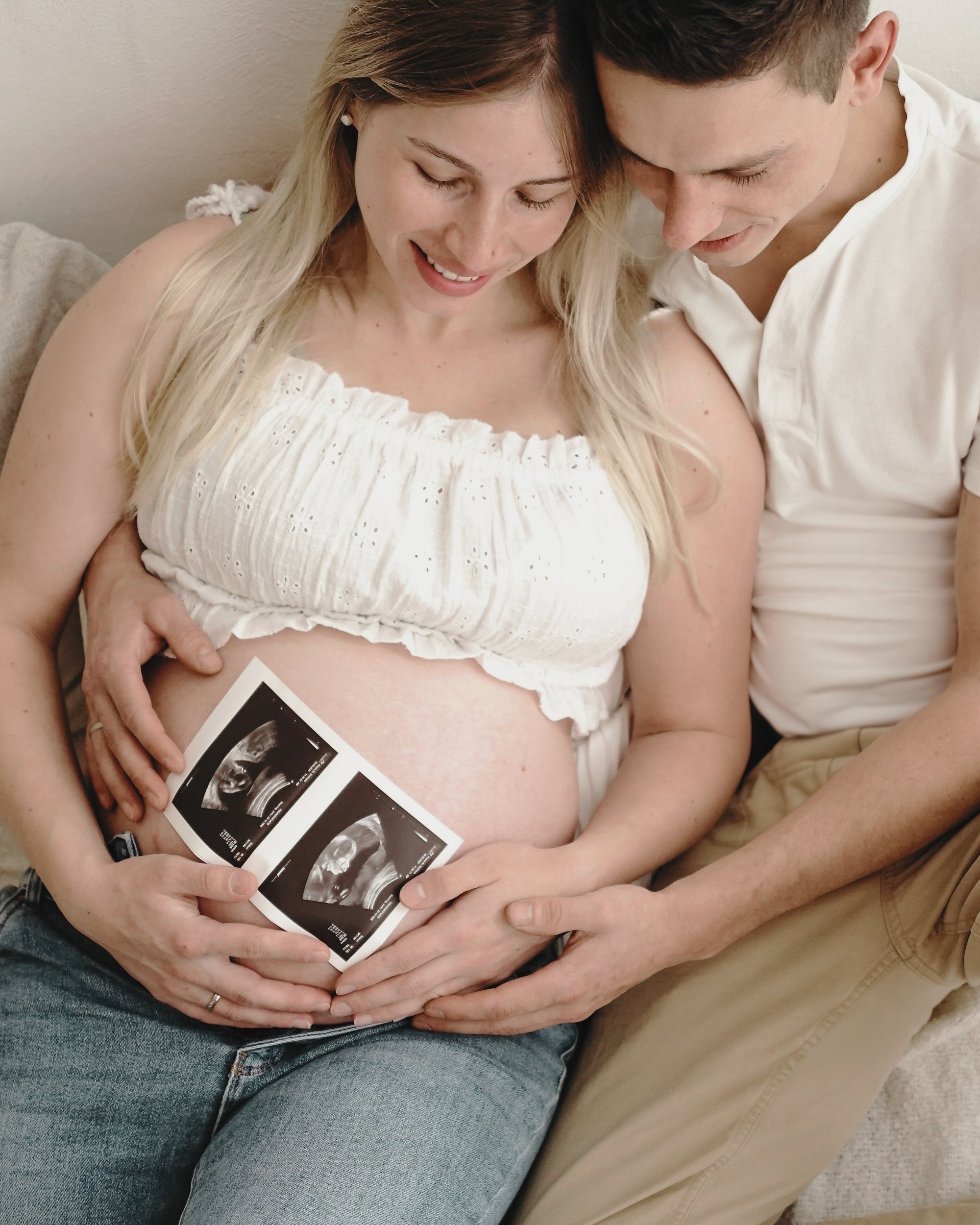 Ein schwangertes Paar sitzt zusammen auf einer Couch, die Frau hält Ultraschallbilder ihres Babys. Beide schauen liebevoll auf den Bauch.
