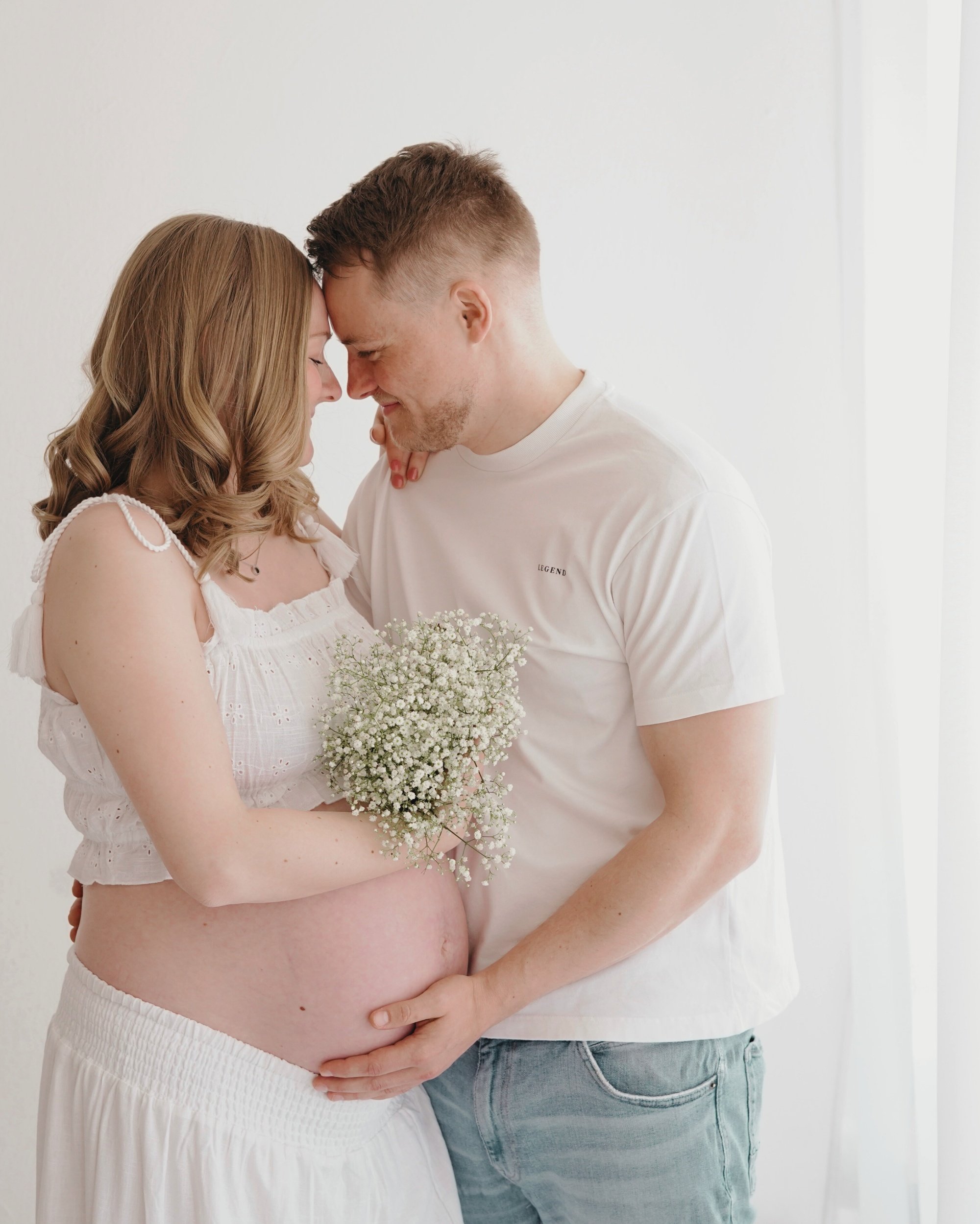 Ein schwangertes Paar steht im weißen Zimmer, berührt sich mit den Stirnen und hält eine Blumenbund