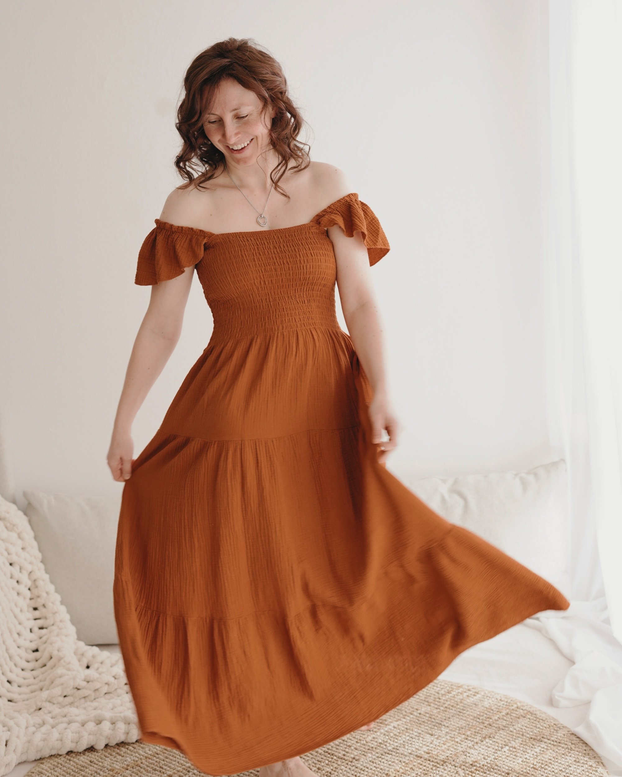Frau in einem orangefarbenen Sommerkleid, die auf einem Sofa mit einer weißen Decke sitzt, lächelnd und das Kleid schwingend.