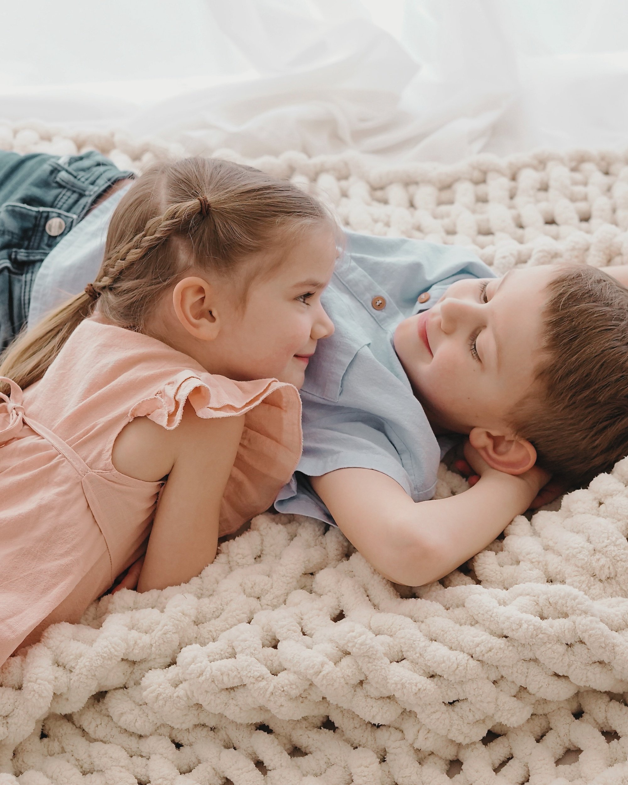 Zwei Kinder, ein Mädchen und ein Junge, liegen sich lachend auf einem weichen weißen Teppich. Sie schauen sich an und scheinen Spaß zu haben.