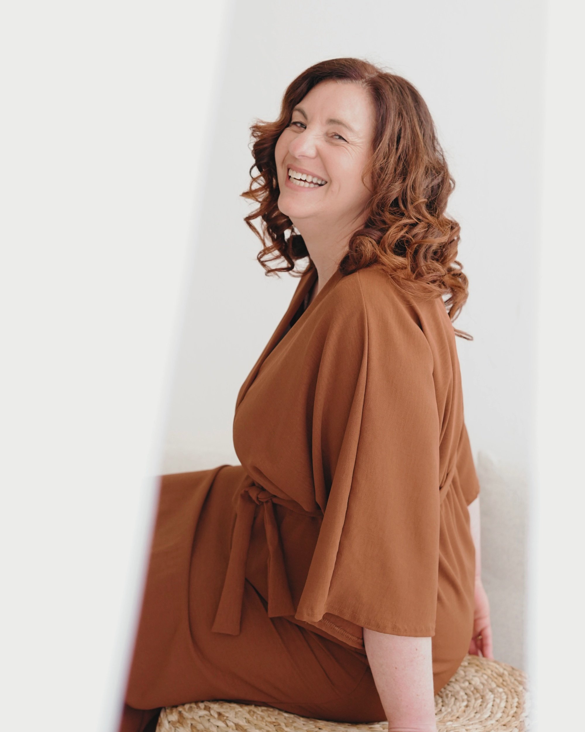 Eine lachende Frau mit lockigem braunem Haar trägt ein braunes Kleid und sitzt auf einem geflochtenen Sitzmöbel.