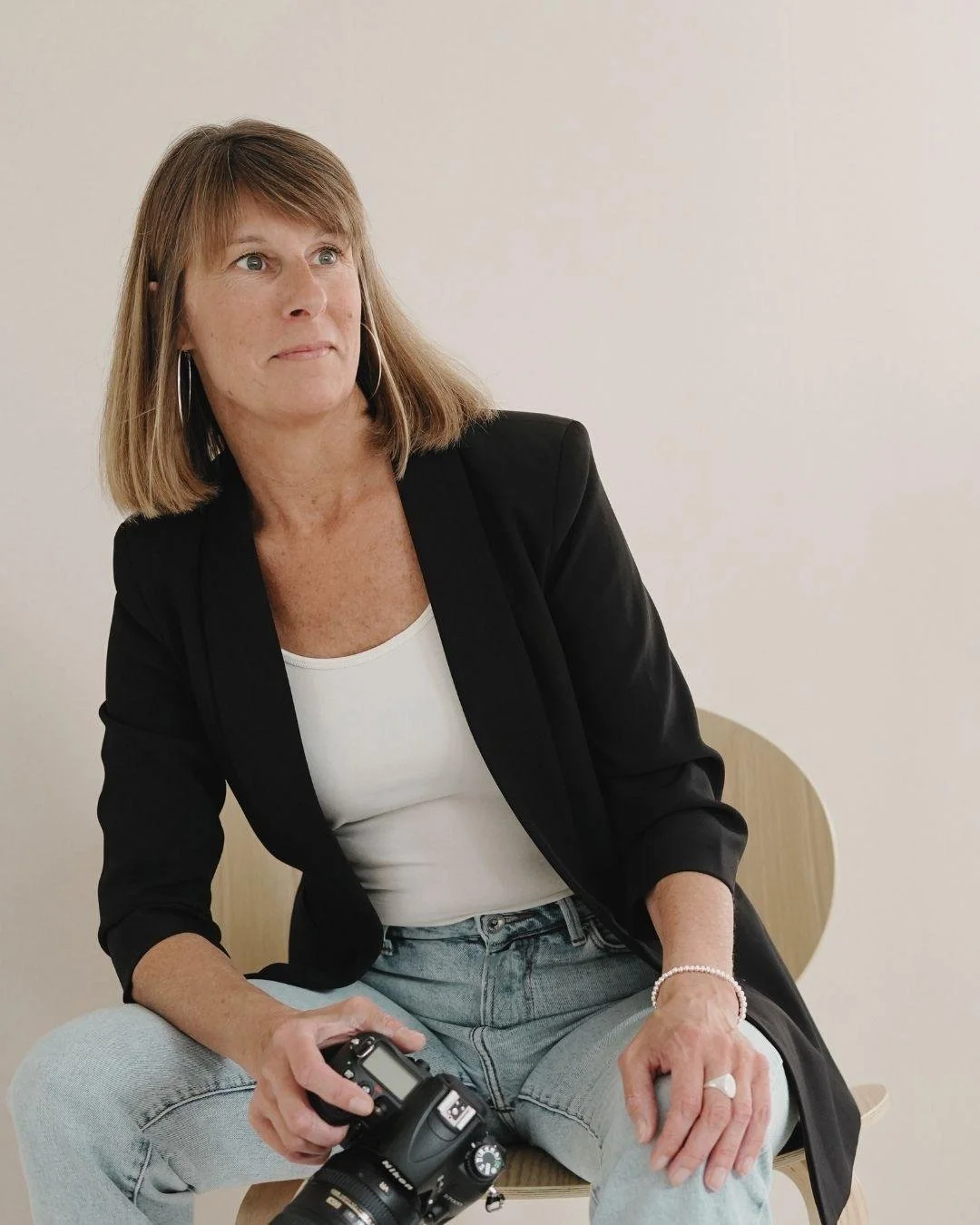 Eine Frau sitzt auf einem Stuhl, trägt ein schwarzes Jackett, eine weiße Bluse, Jeans, hält eine Kamera in ihrer Hand und blickt nach links.