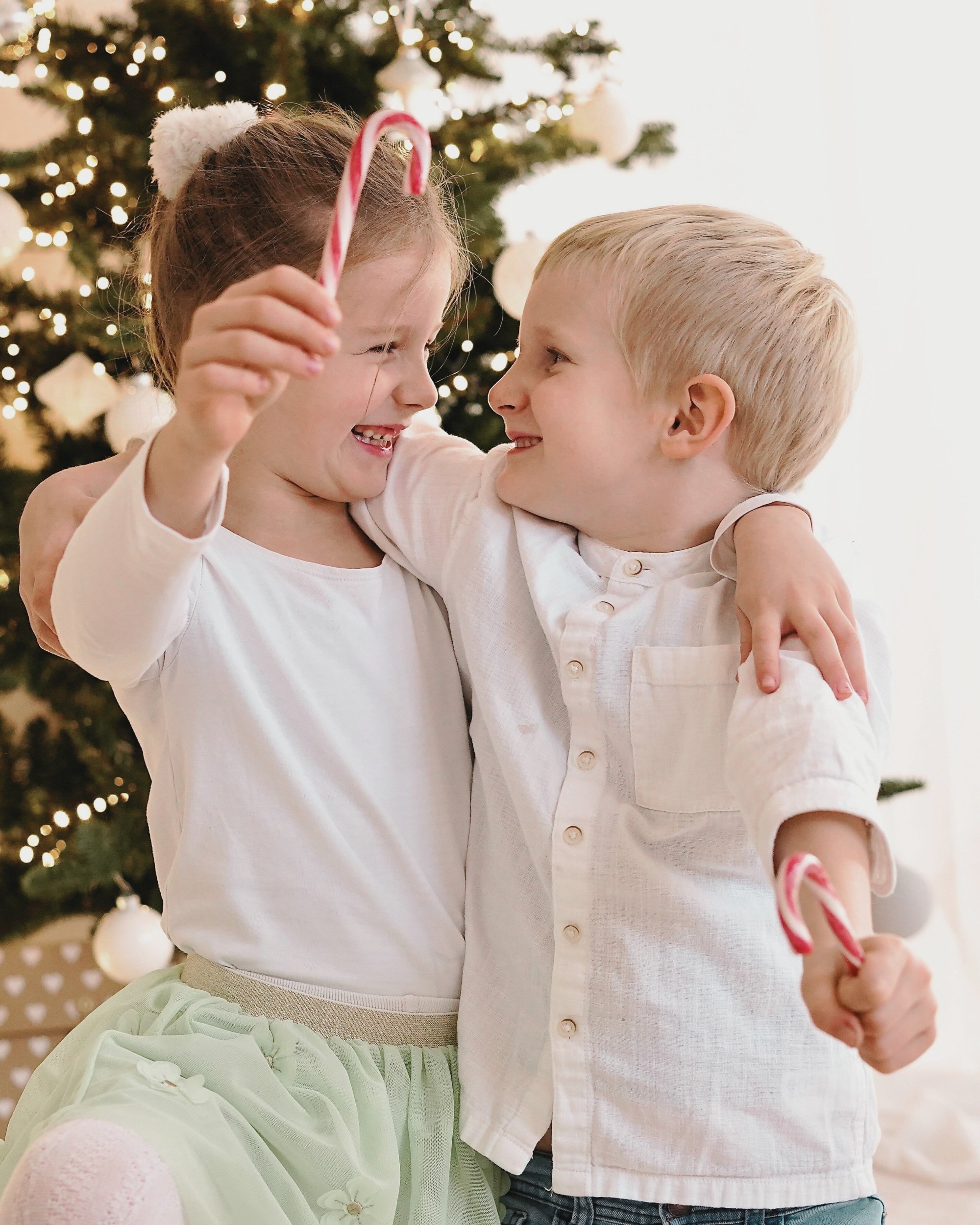 Zwei Kinder, ein Mädchen und ein Junge, freuen sich, während sie sich umarmen, vor einem geschmückten Weihnachtsbaum, das Mädchen hält eine Zuckerstange in der Hand.
