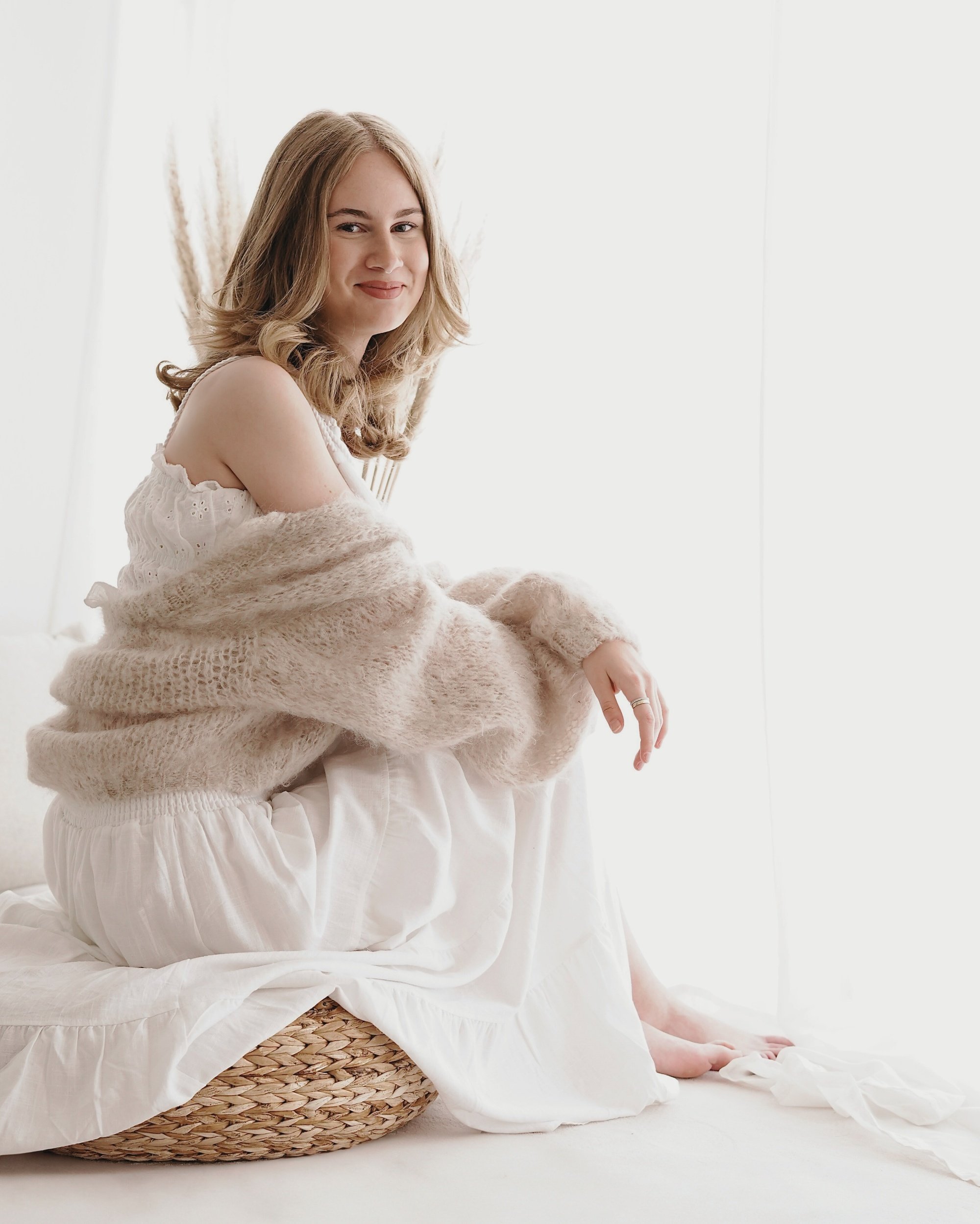 Junge Frau mit grauen Haaren in weißen Kleid, sitzend auf einem Fußhocker, umgeben von natürlichem Licht, trägt einen lockeren Pullover und schaut lächelnd in die Kamera.