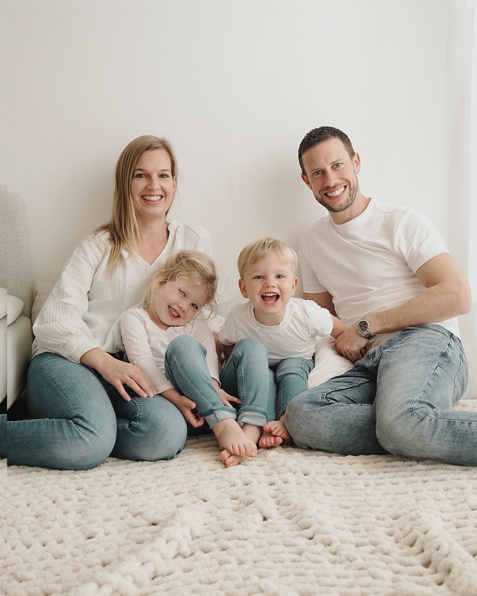 Familie mit vier Personen, zwei Kinder und zwei Erwachsene, sitzt gemeinsam auf einem Teppich vor einer weißen Wand, lachend und happy.