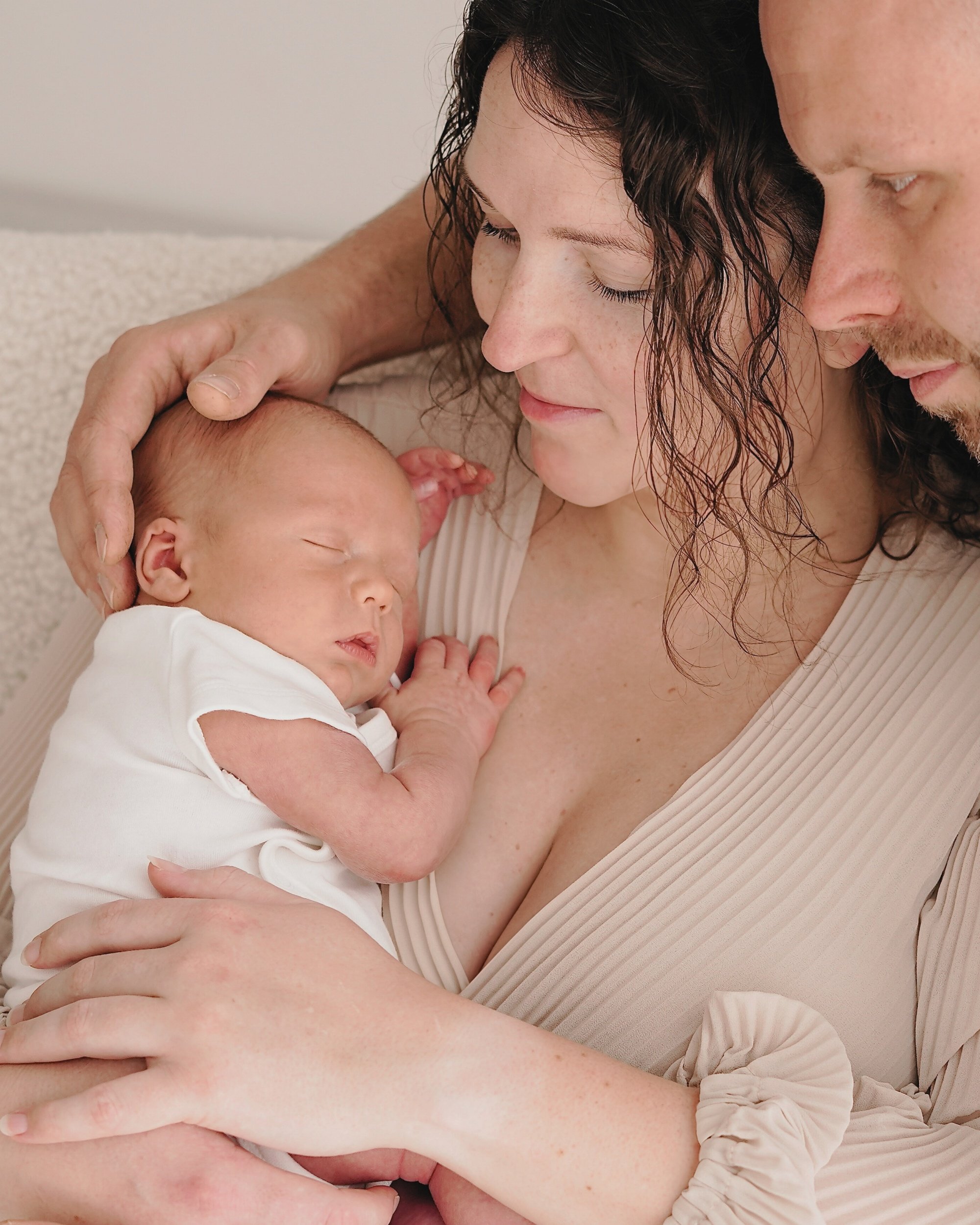 Ein Baby wird von einer Frau gehalten, während ein Mann daneben steht. Alle schauen das schlafende Baby liebevoll an.