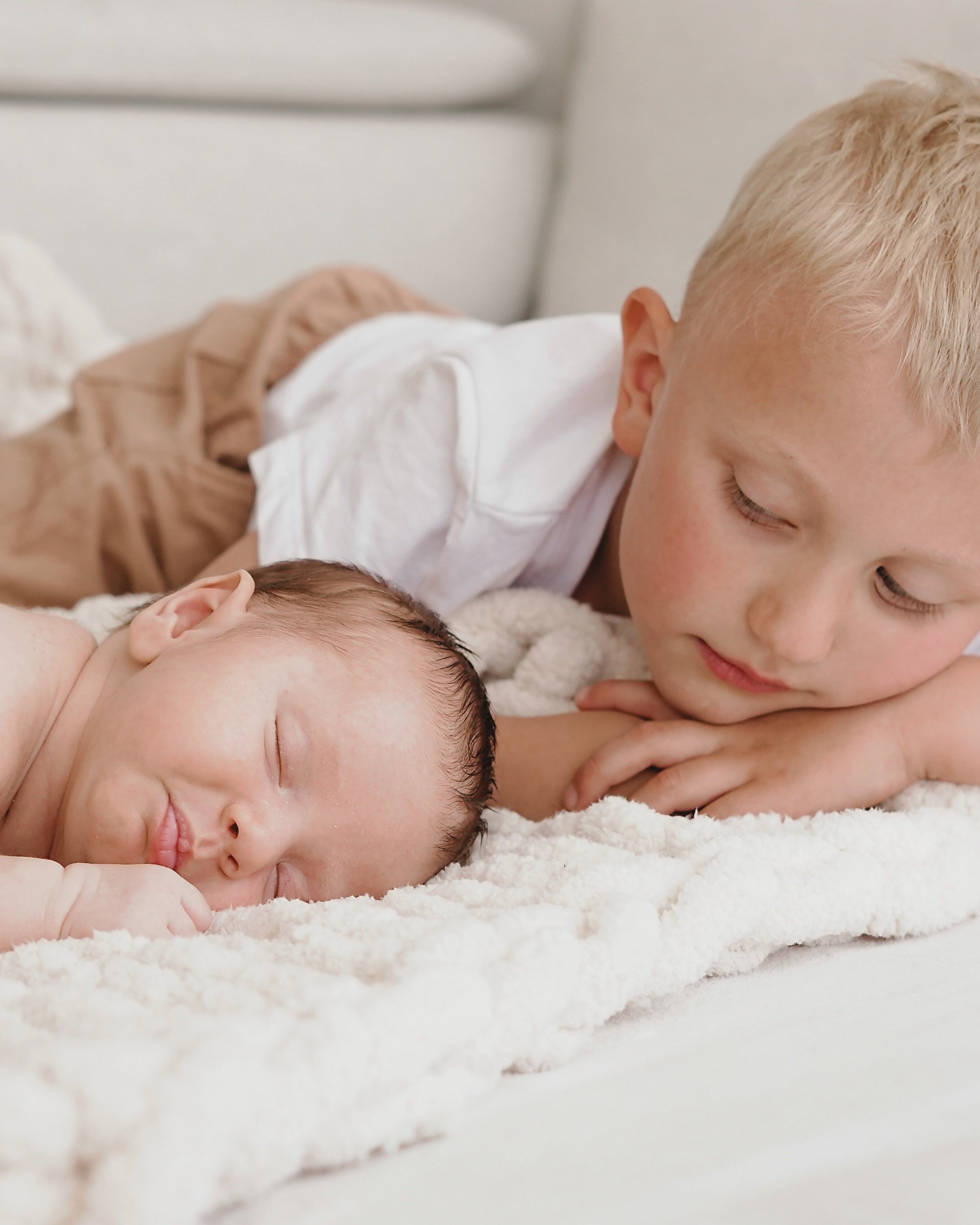 Ein älterer Junge liegt auf einem Bett und schaut auf ein schlafendes Baby, beide sind auf einem weißen Bett mit weicher Decke.