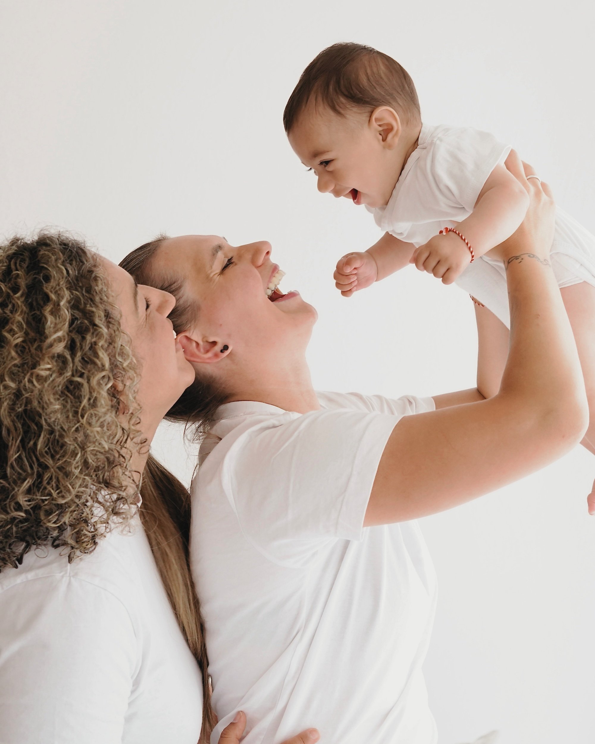 Drei Frauen, zwei mit Locken, eine mit glatten Haaren, lachen und freuen sich, während eine der Frauen ein Baby hochhebt, das lächelt und nach ihnen greift.