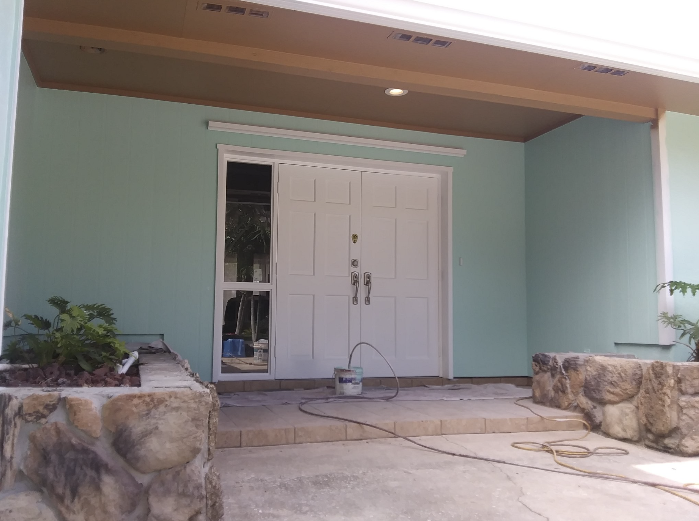 Front porch with white double doors, surrounded by light teal walls, stone planters with plants, and construction tools including a paint bucket and extension cord.