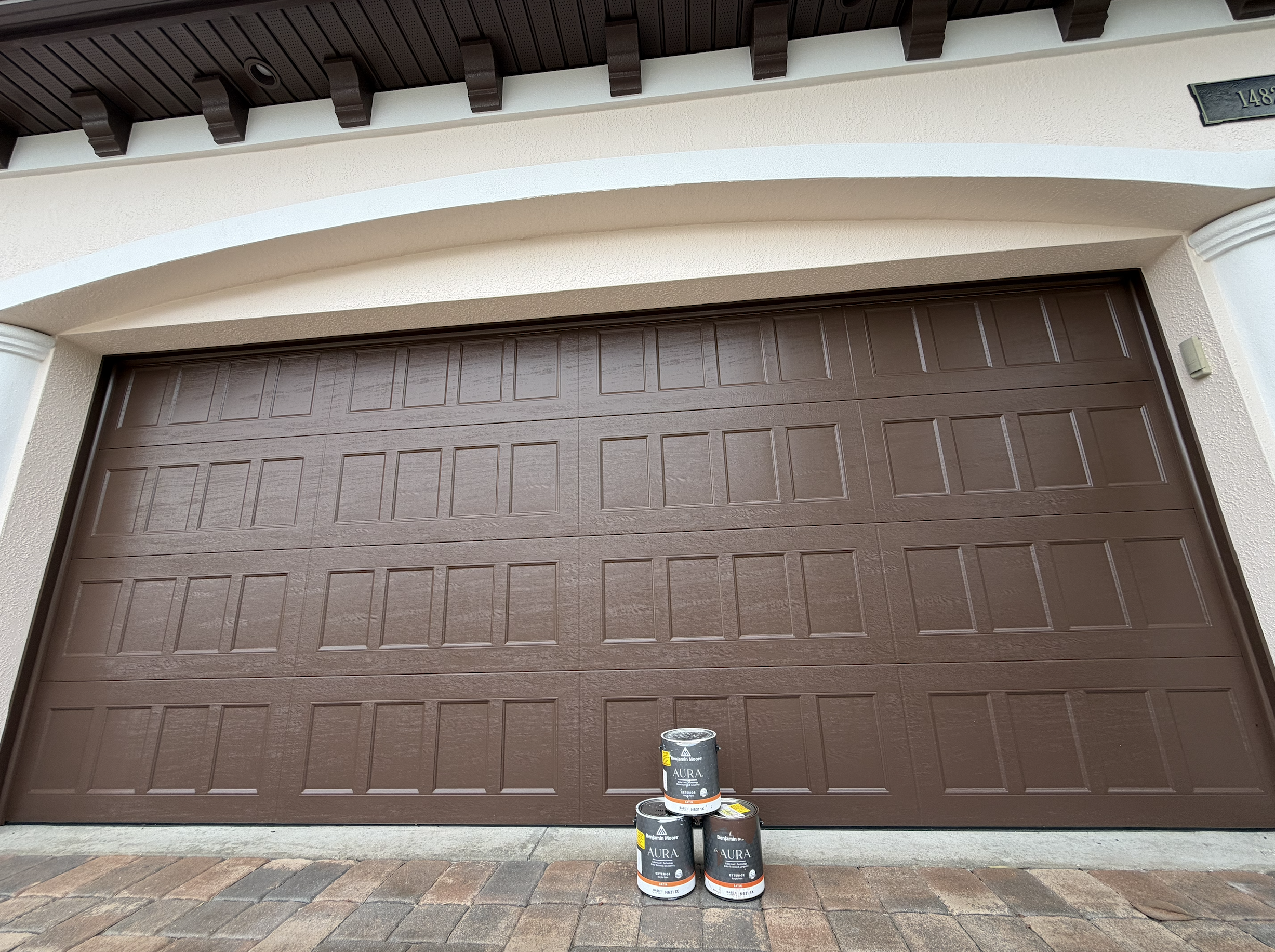 A closed brown garage door with paint cans and a paint roller in front, indicating ongoing painting work.
