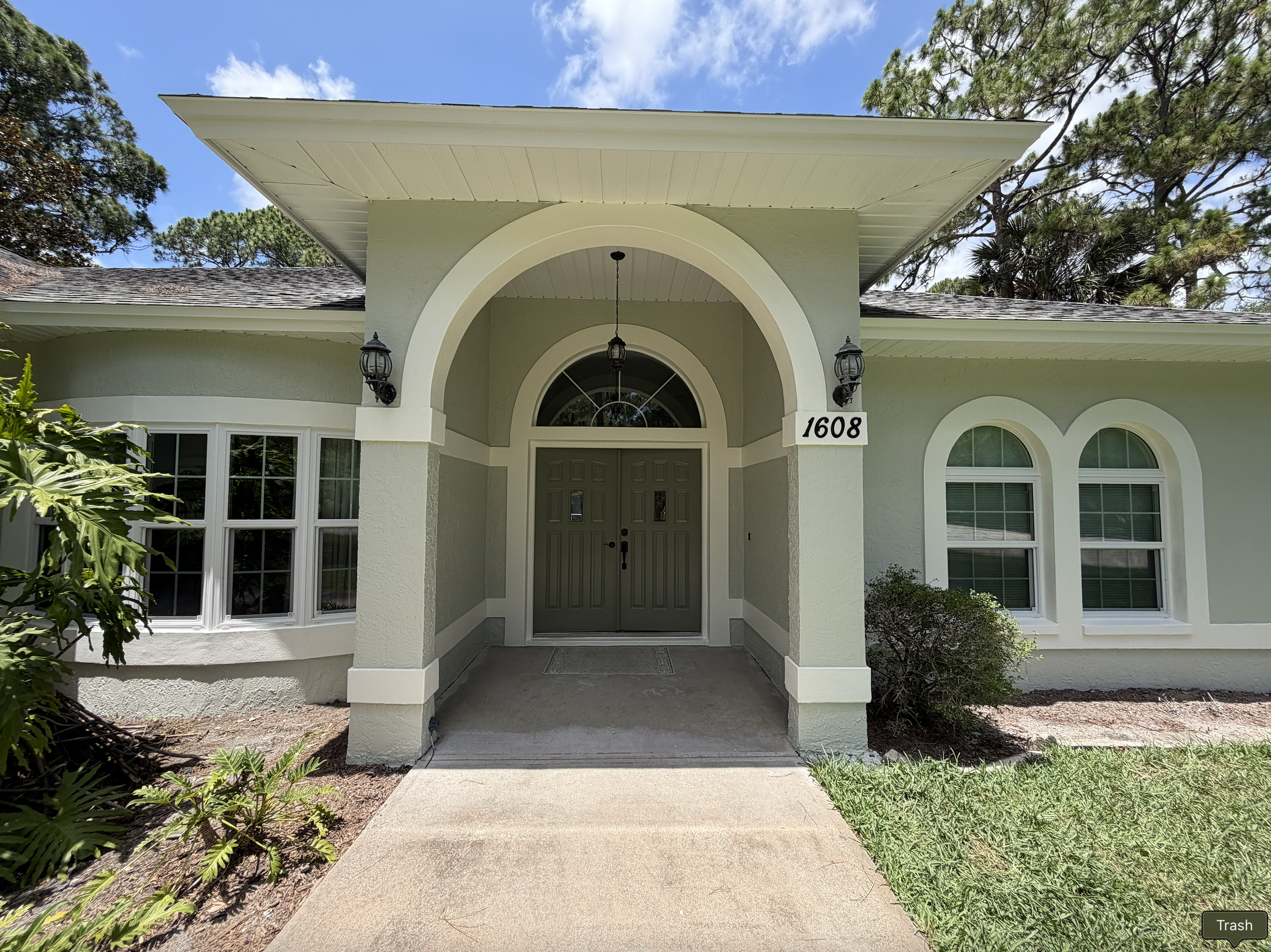 Front view of a light green house with a covered porch, arched doorway, and the house number 1608 displayed on a plaque. There are black wall-mounted lanterns on either side of the entryway, and trees with green foliage surround the house under a partly cloudy blue sky.