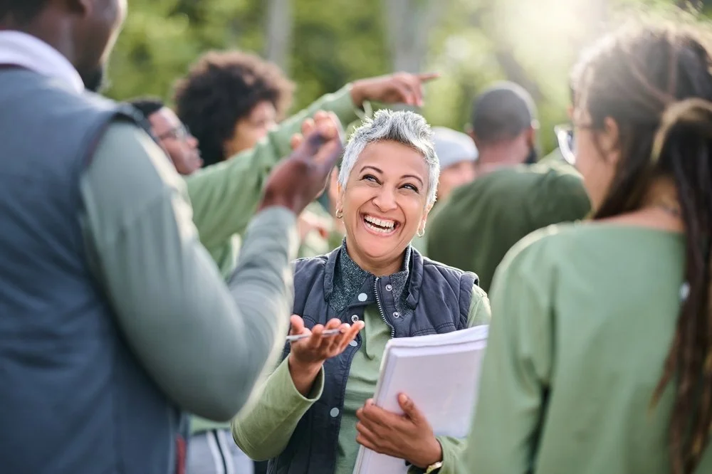 A group of people outdoors, engaging in conversation with a smiling woman holding a notepad, surrounded by trees.