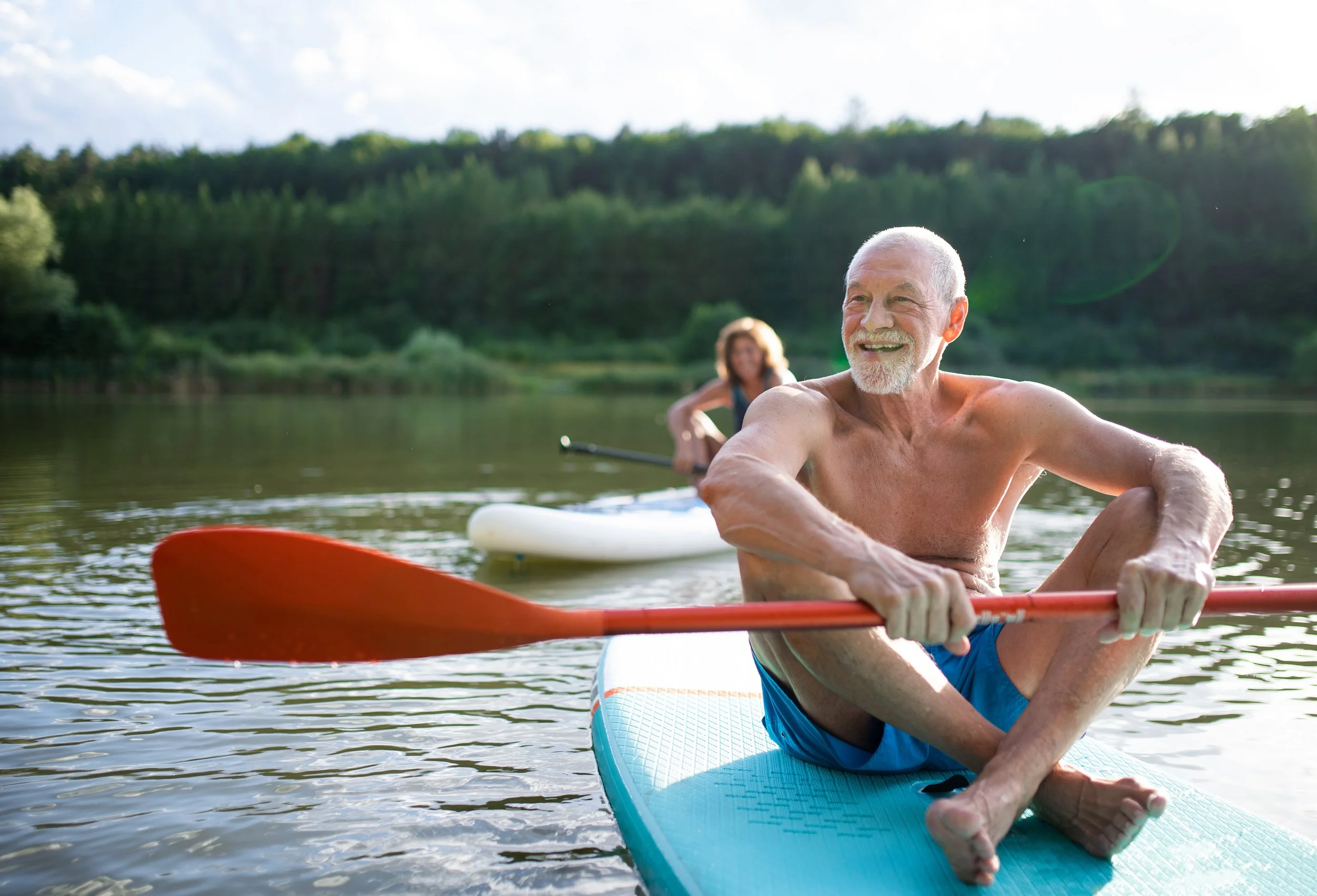 An elderly man with a white beard smiling while sitting on a paddleboard and holding a red paddle on a calm lake, with a woman in the background also paddleboarding.