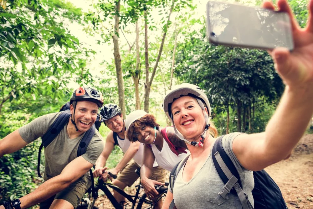 Four friends in riding helmets and casual clothing taking a group selfie while mountain biking on a trail surrounded by green trees.