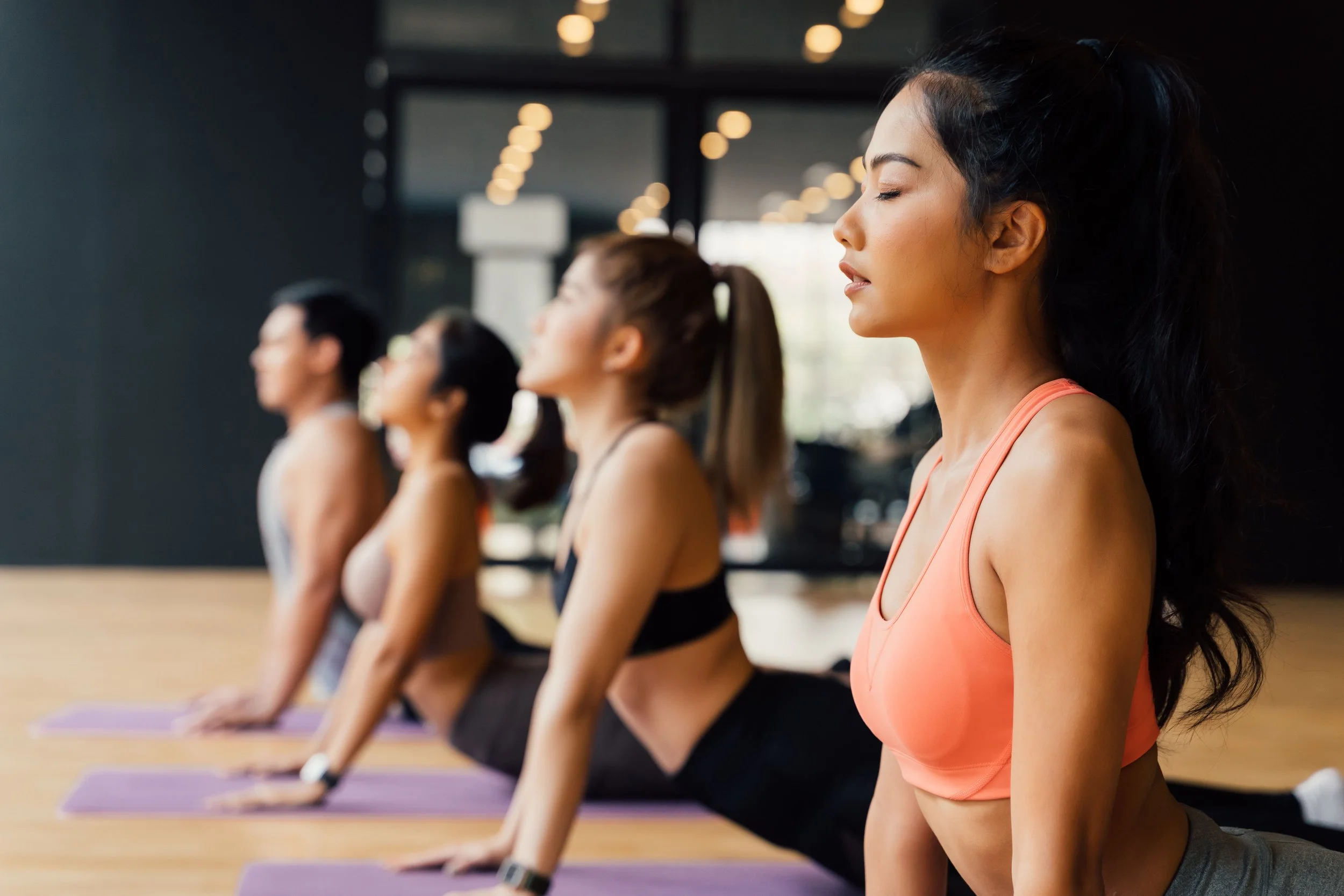 A group of five people practicing yoga in a studio, kneeling on yoga mats with eyes closed and calm expressions.