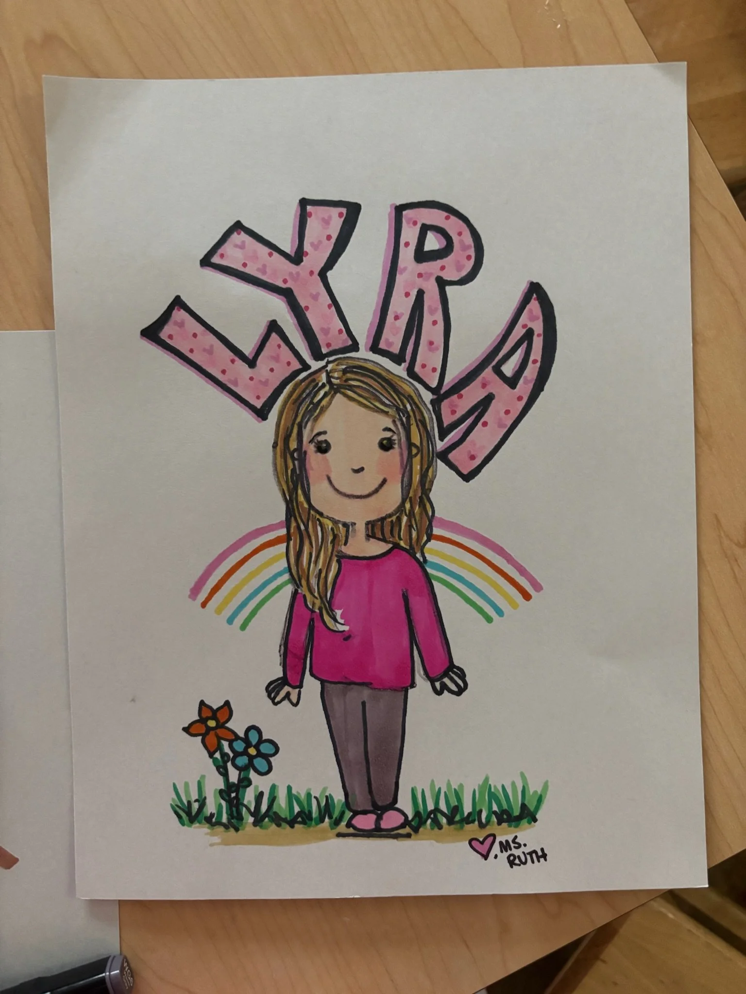 Hand-drawn picture of a smiling girl with long, wavy hair, wearing a pink shirt and gray pants, standing on grass with flowers and a rainbow in the background. The name 'Lyra' is written above her in large, pink, decorated letters, and there is a sma