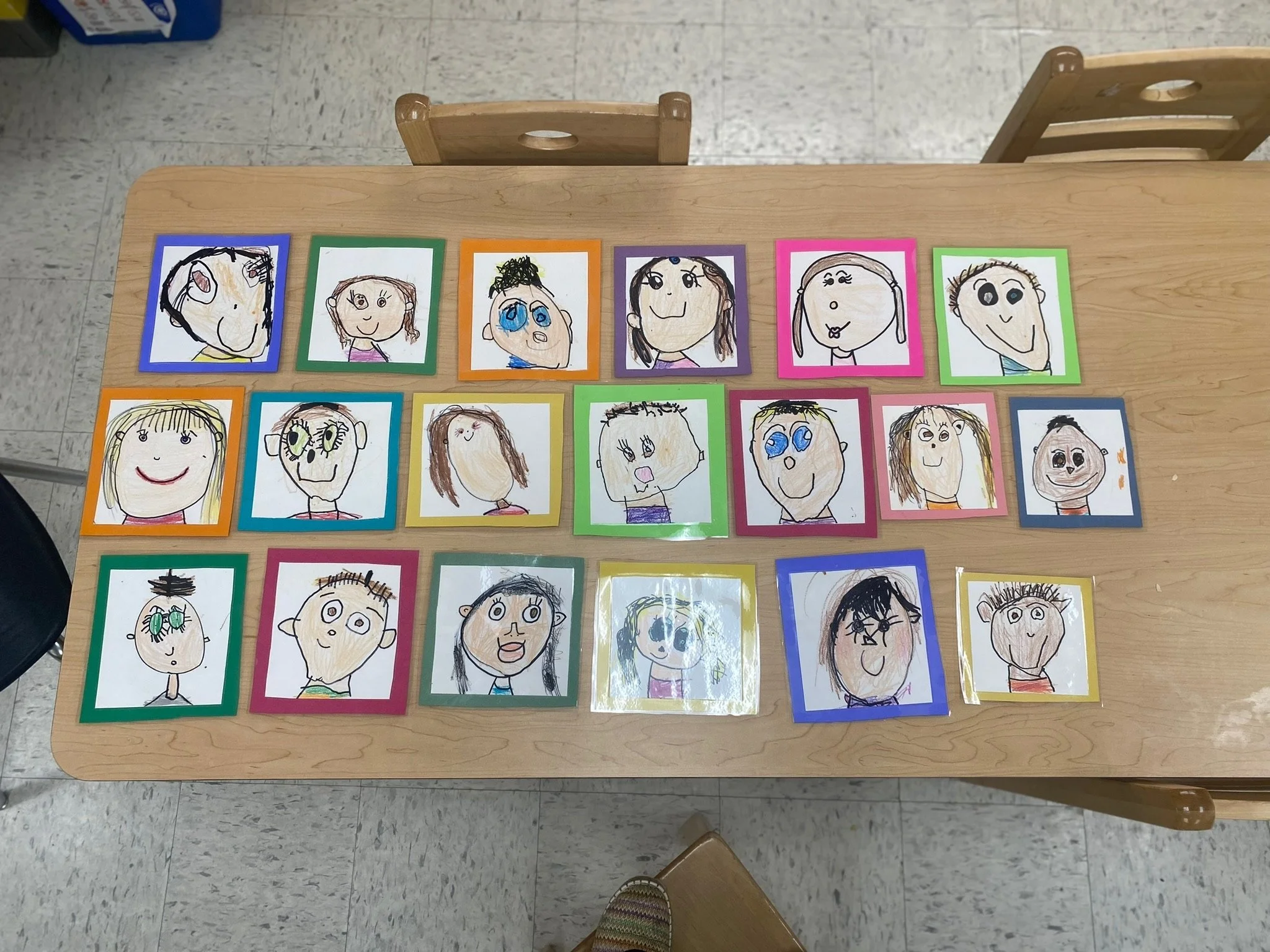 Collection of children's drawings of faces, each framed with colorful borders, arranged on a wooden table.