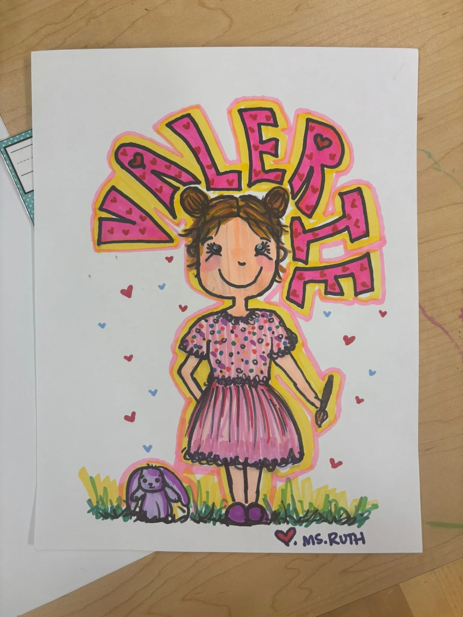A colorful illustration of a girl with brown hair in buns, smiling, wearing a pink polka dot dress, holding a marker, with the name 'Vanelie' in large pink and yellow letters above her. A purple bunny sits on the grass near her. The drawing is signed