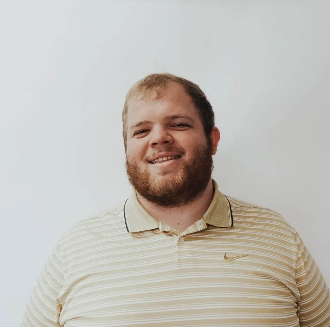 A man with a beard and short hair wearing a cream and beige striped polo shirt, smiling, standing against a plain white wall.