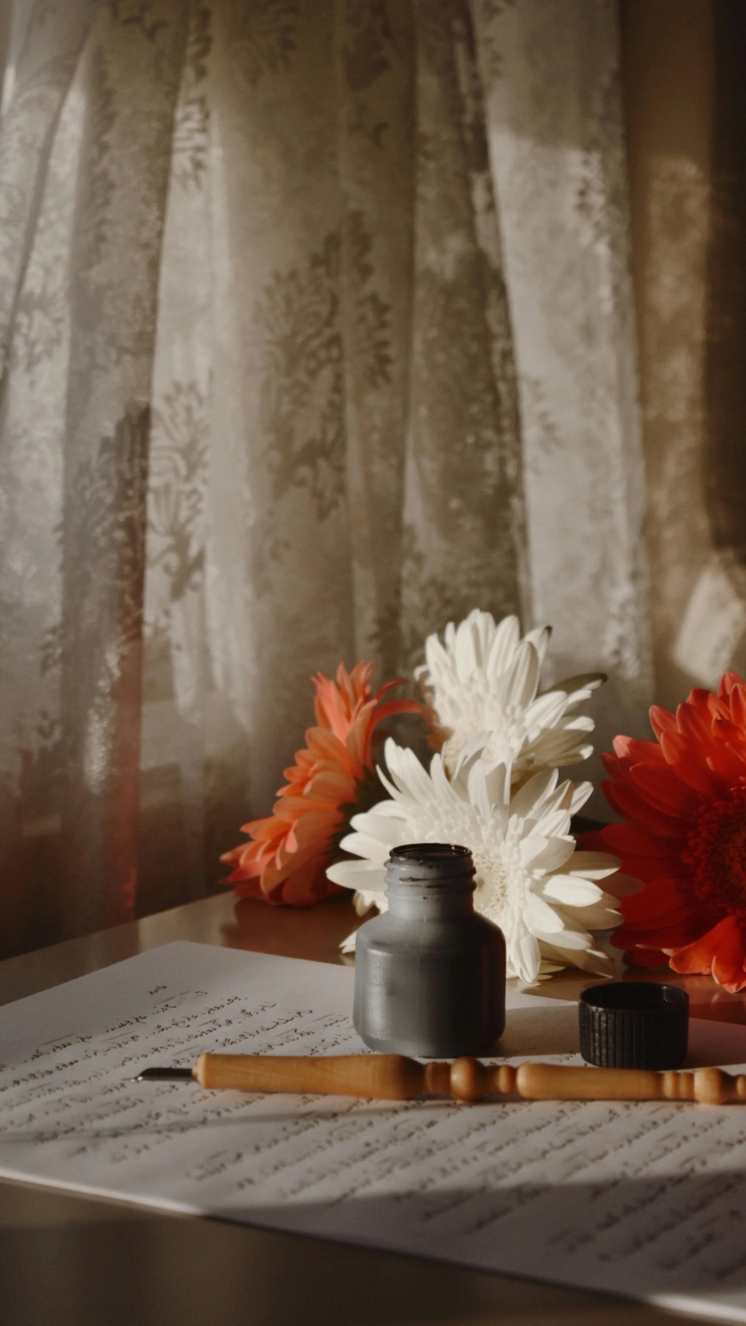 A handwritten note on a table beside a pen and flowers, arranged near a softly lit window with a sheer curtain.