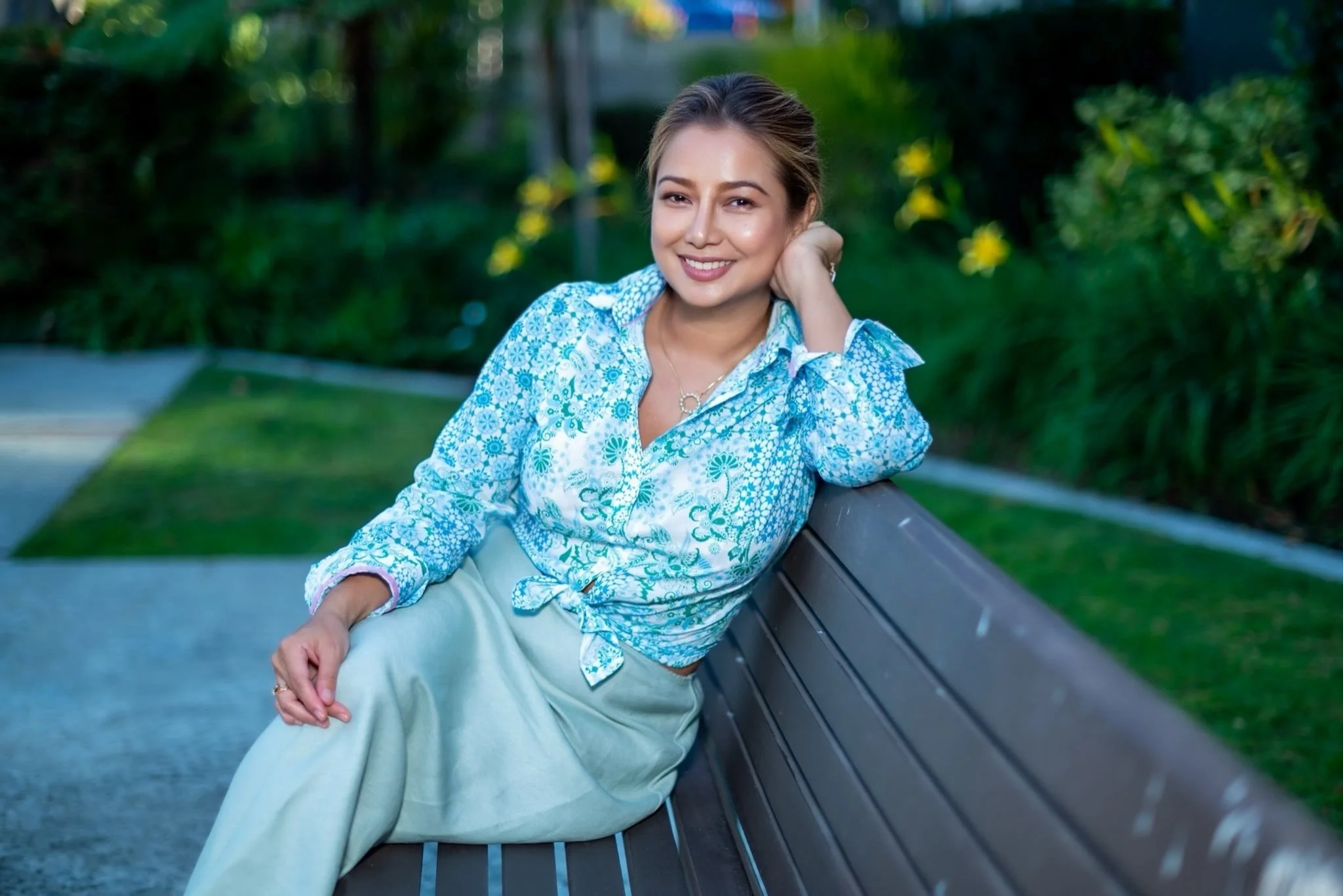Young woman sitting on a park bench, resting her head on her hand, surrounded by soft greenery — evoking calm reflection and emotional openness.
