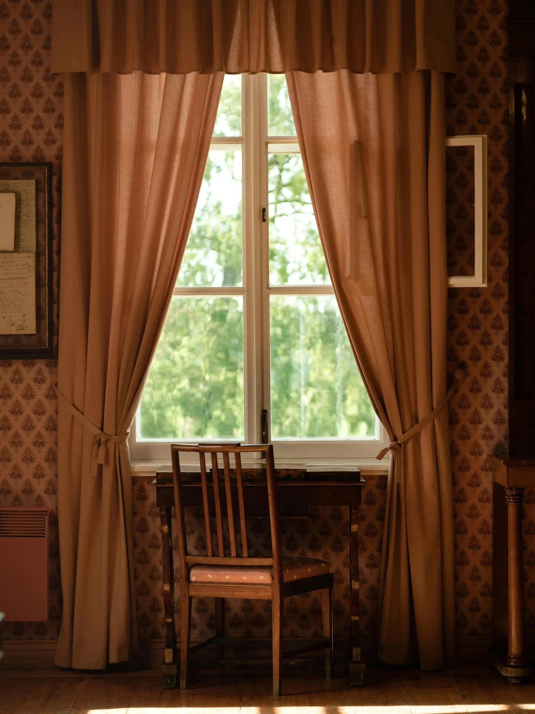A wooden chair facing a window with soft daylight filtering through curtains, suggesting quiet waiting and reflection.
