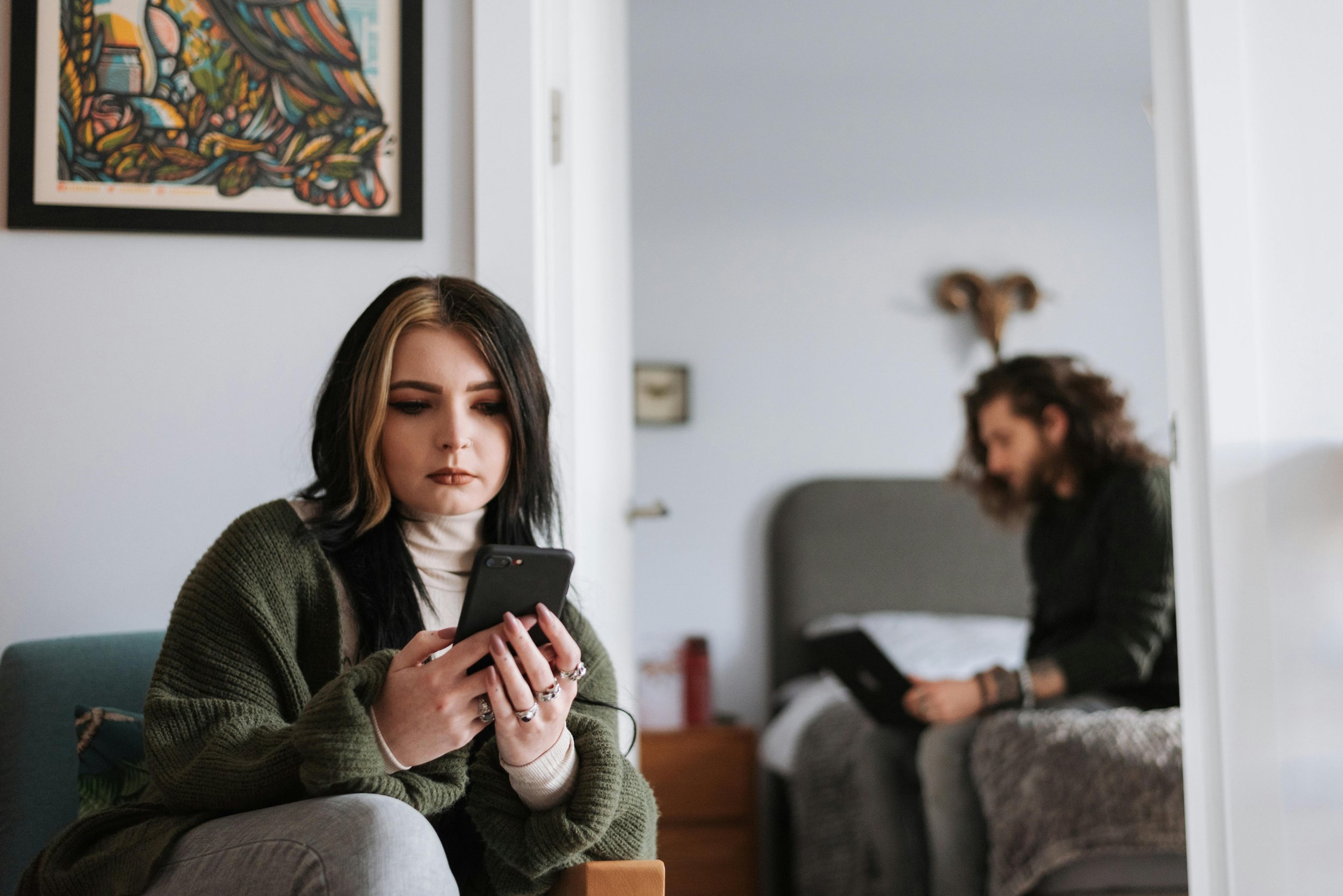 Couple sitting separately in the same room, woman using a phone in the foreground while her partner works on a laptop in the background